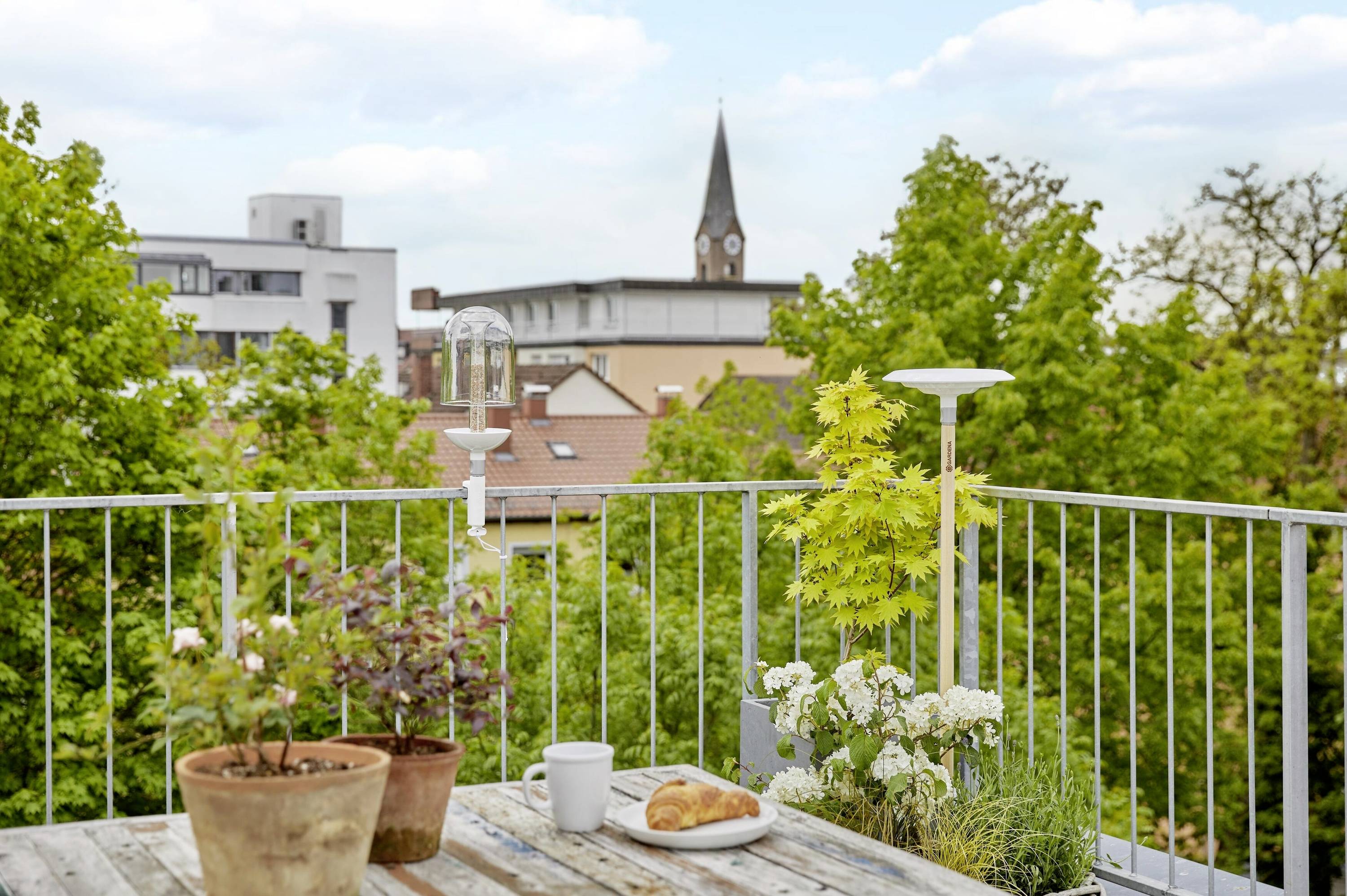 Taras zTablem, roślinami, filiżanką kawy i croissantem. W tle widać drzewa i wieżę kościelną.