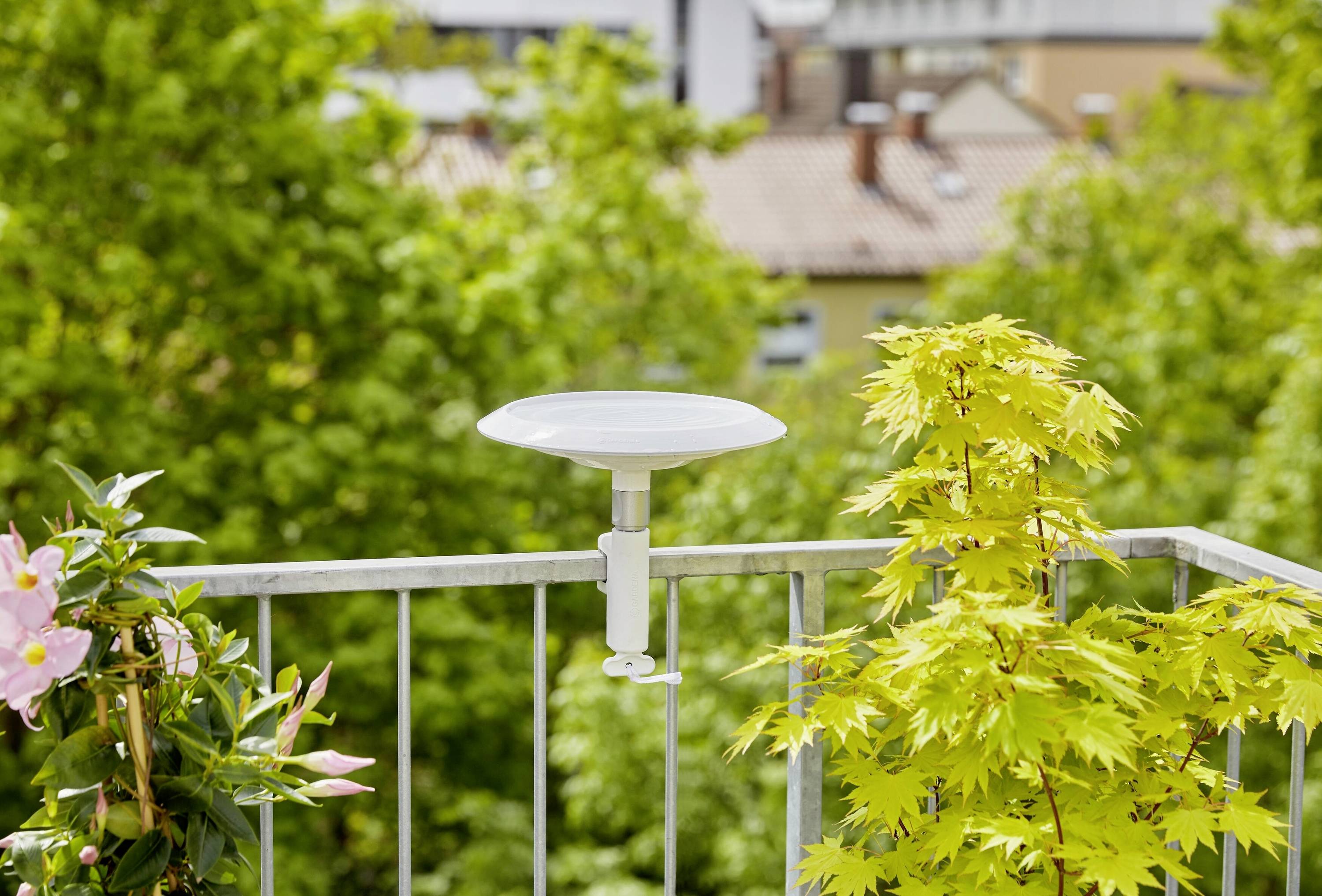 Biała antena satelitarna jest zamontowana na balustradzie balkonu, otoczona zielonymi roślinami, z krajobrazem miasta w tle.