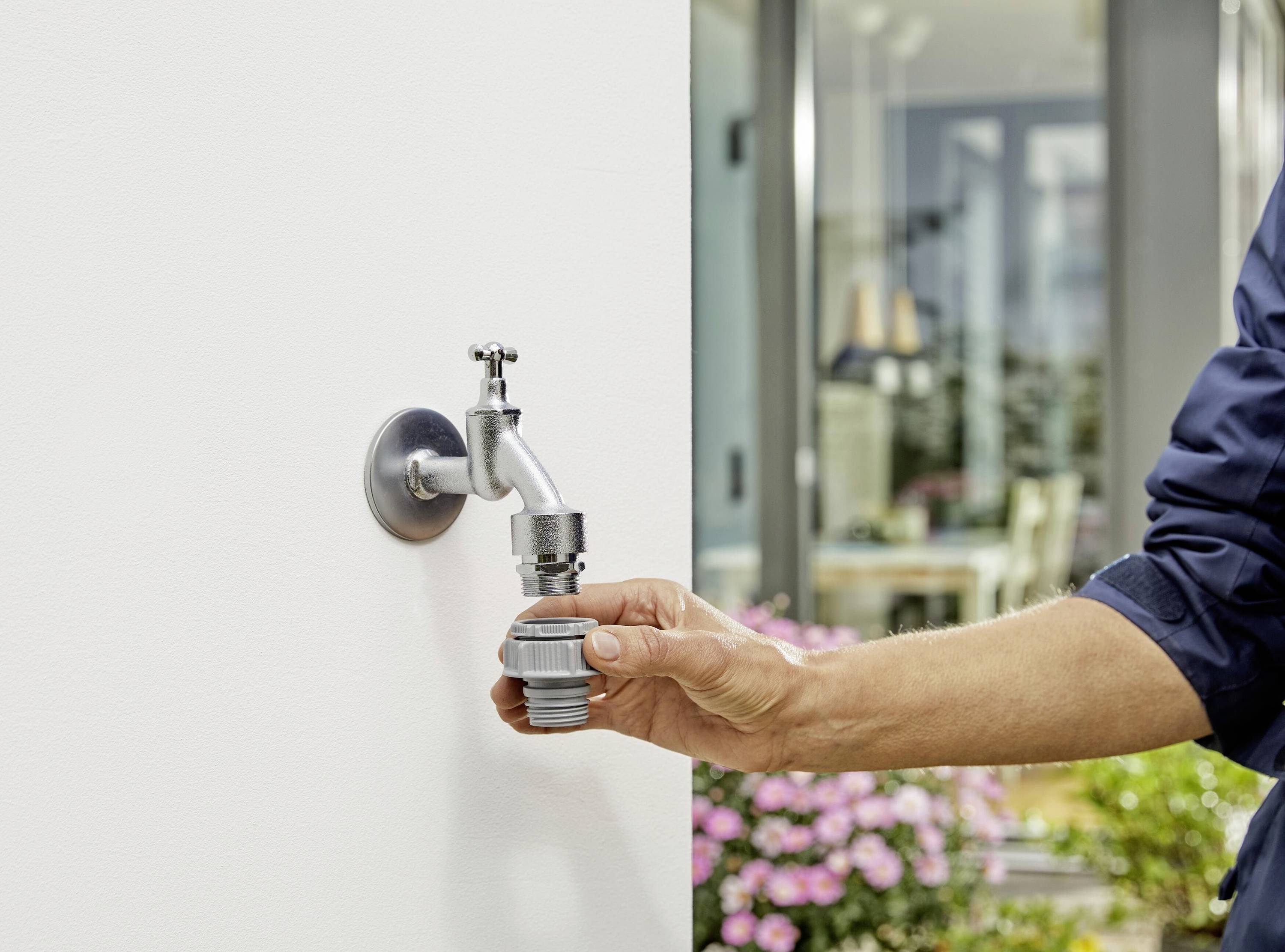Osoba podłącza wąż ogrodowy do外ętrznego kranu na białej ścianie, z domem i rabatą kwiatową w tle.