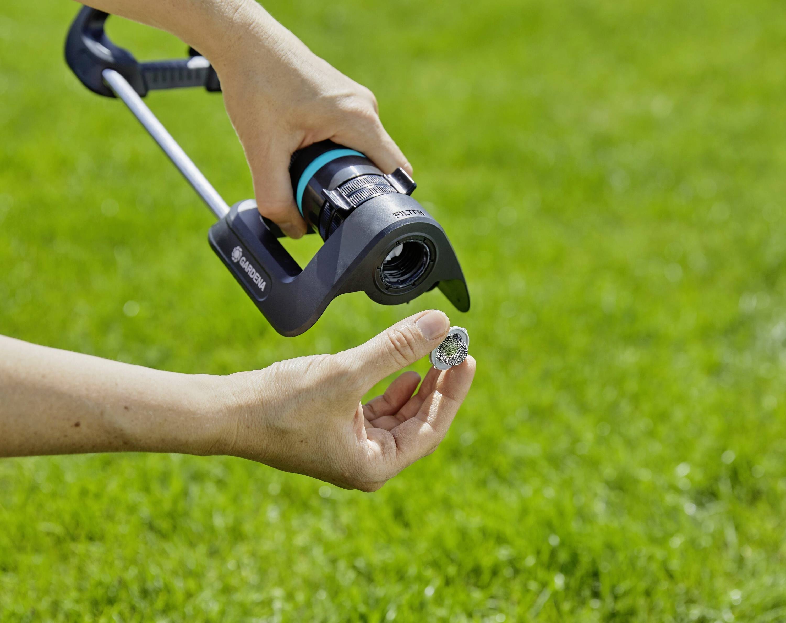 Osoba trzyma głowicę zraszacza kołowego i reguluje dyszę monetą nad zieloną trawą.