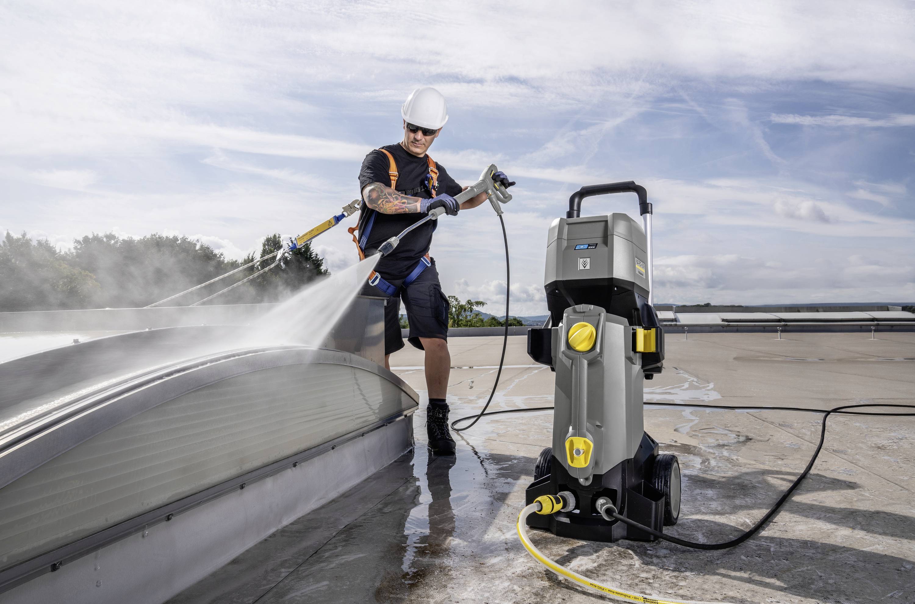 Osoba czyści dach myjką wysokociśnieniową, otoczona błękitnym niebem i chmurami. Nosi prawidłowe ubranie ochronne.