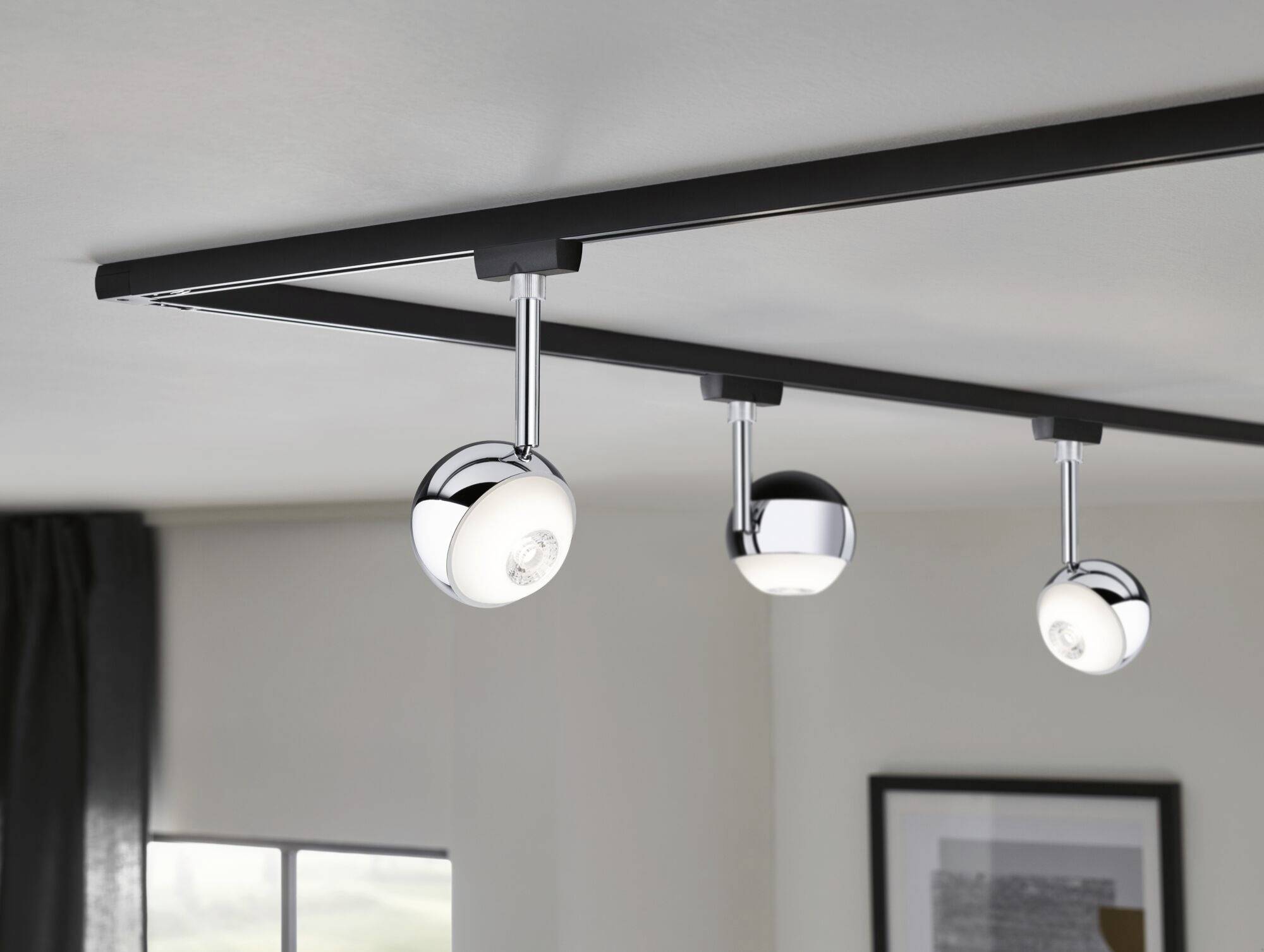 Trzy chromowane reflektory szynowe na suficie oświetlają nowoczesny, minimalistycznie urządzony pokój z jednym oknem i obrazem.