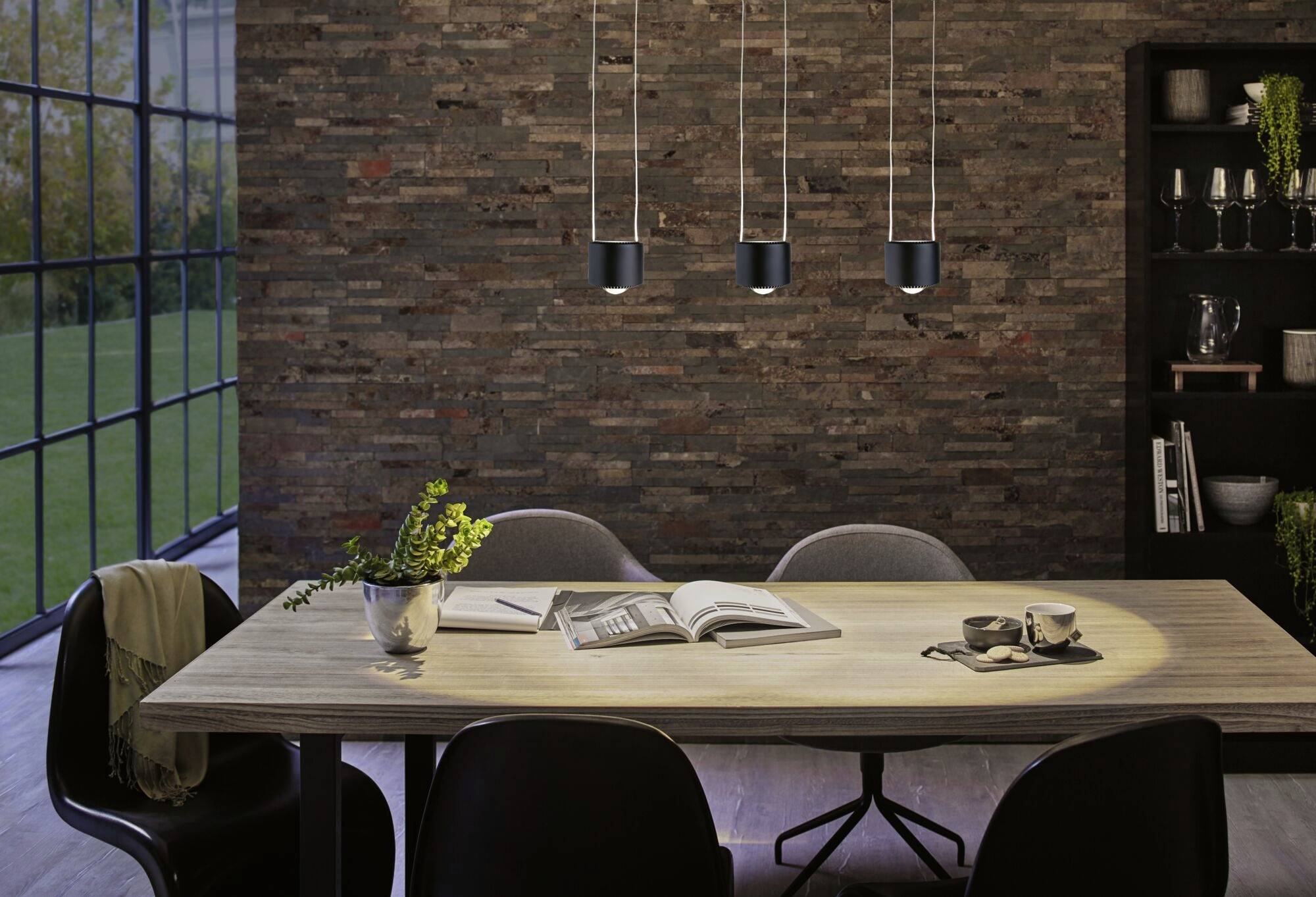 Nowoczesna jadalnia z drewnianym krzesłem, stołem, trzema wiszącymi lampami i ścianą z cegły. Otwarta książka i rośliny na stole.