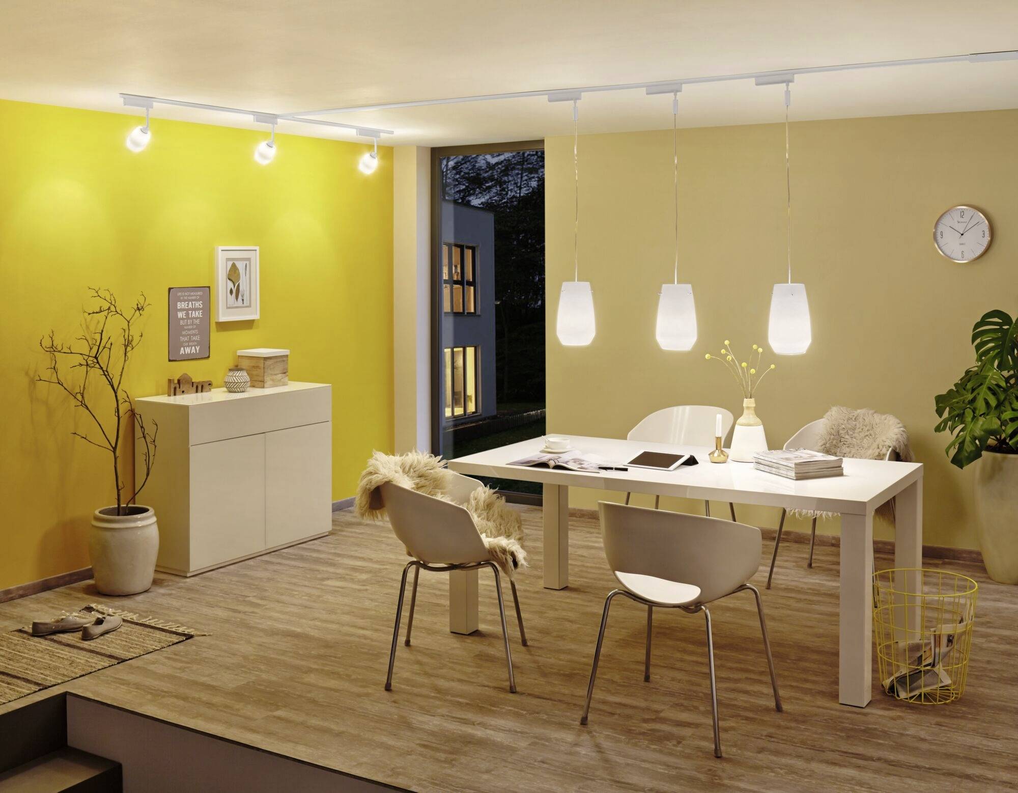Jadalnia z żółtą ścianą, stołem jadalnianym i białymi krzesłami. Lampy wiszące nad stołem. Okno ukazuje widok na wieczorową przestrzeń na zewnątrz.
