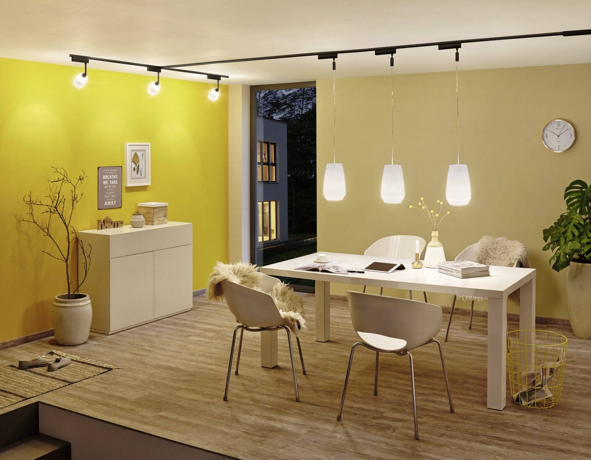 Nowoczesny salon z żółtymi ścianami, białym stołem z czterema krzesłami, wiszącymi lampami oraz przeszkloną, otwartą bramą prowadzącą na balkon.