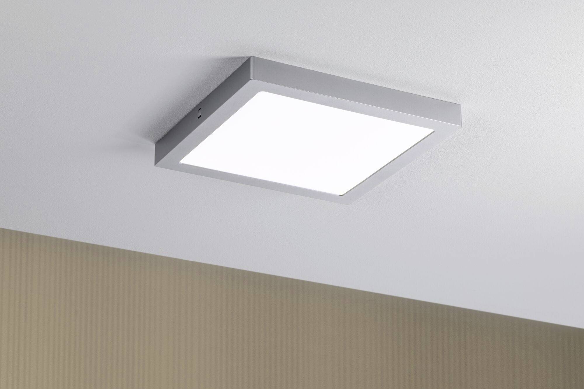 Lampa sufitowa o kwadratowym, nowoczesnym wzornictwie, zapewniająca równomierne oświetlenie. Zamocowana na białym suficie nad beżową ścianą.