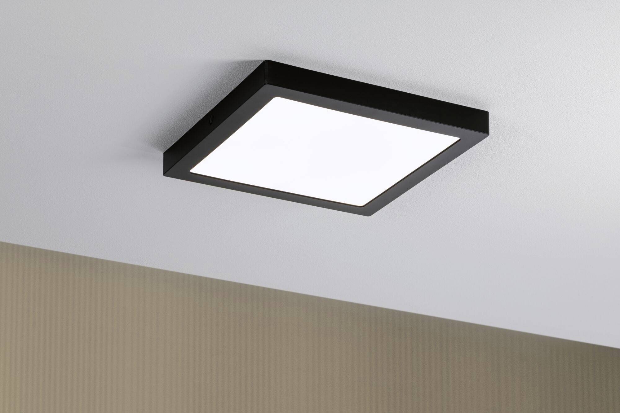 Kwadratowa lampa sufitowa z czarną ramą na białym suficie. Świeci jasno i równomiernie.
