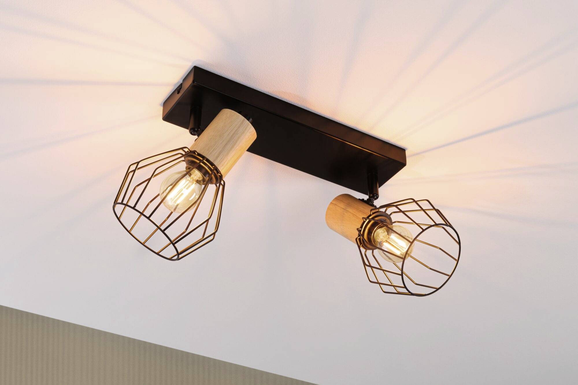 Dwie lampy sufitowe z drewnianymi mocowaniami i metalowymi osłonami w formie siatki, które rozpraszają ciepłe światło we współczesnym wnętrzu.
