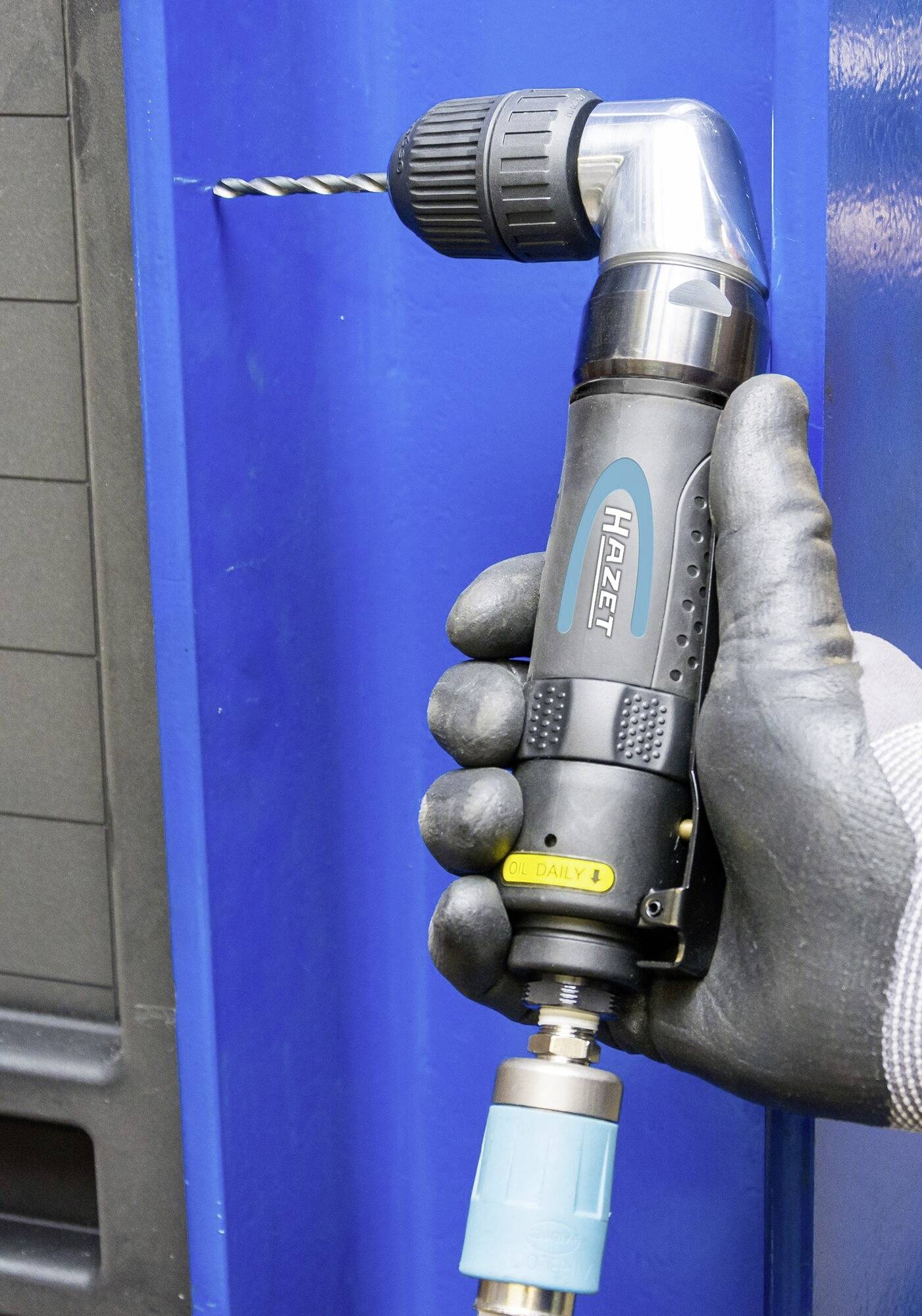 Ręka trzyma pneumatyczny klucz kątowy, który wierci w niebieskim elemencie metalowym. Klucz posiada czarne, ergonomiczne wzornictwo uchwytu.