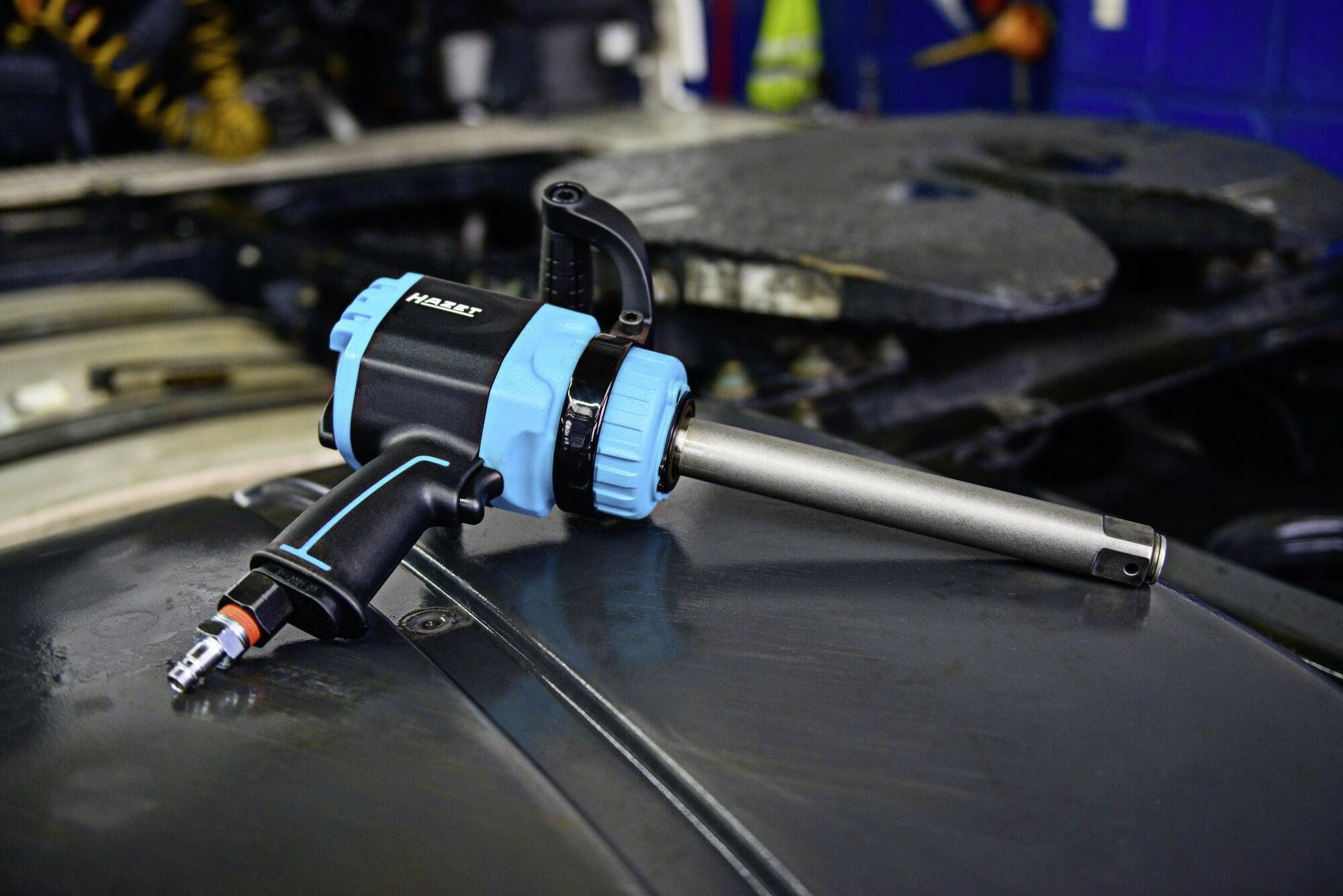 Niebieski i czarny klucz udarowy leży na metalowej powierzchni w warsztacie. W tle widoczne są części samochodowe.