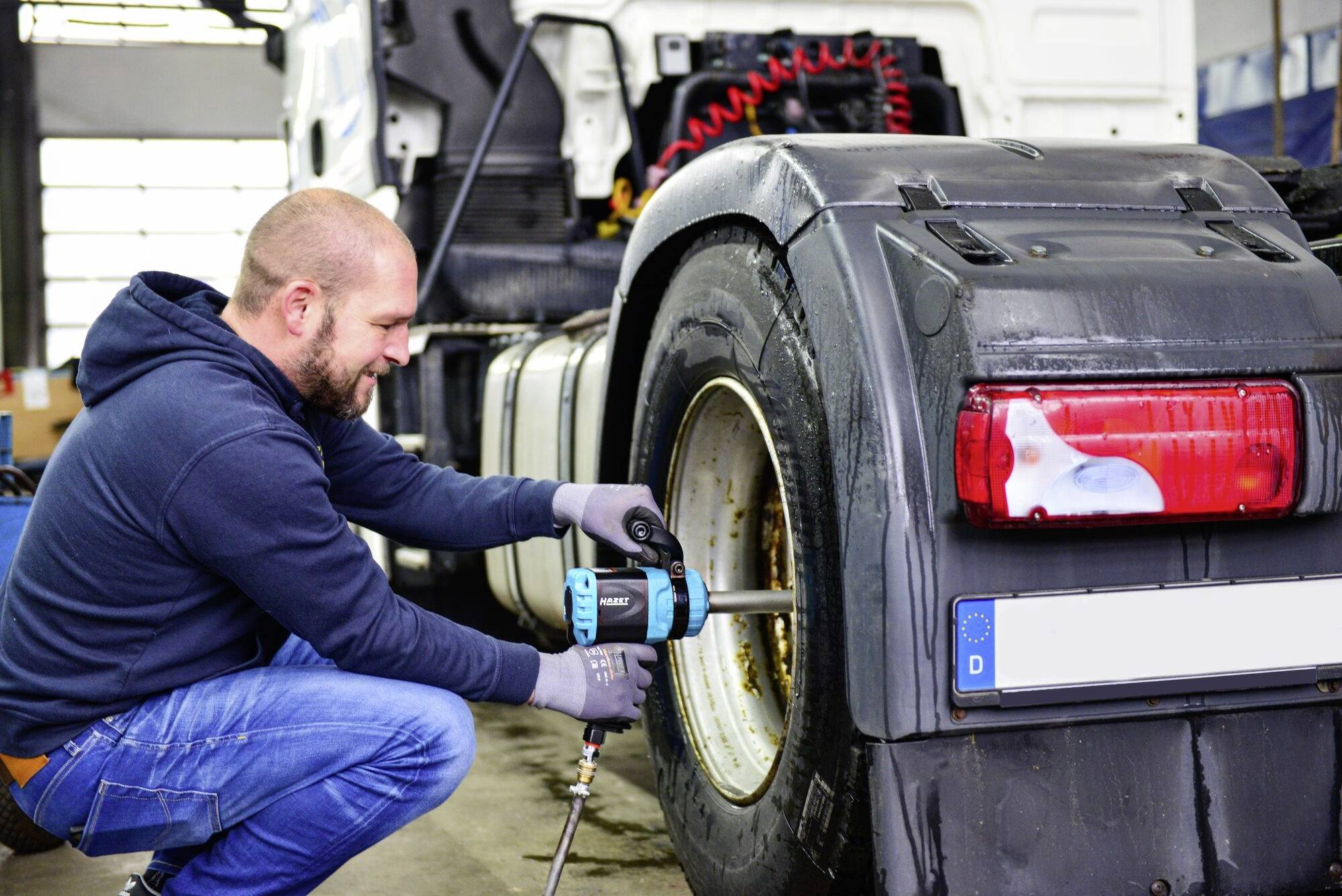 Mężczyzna naprawia oponę ciężarówki za pomocą elektronarzędzia. Ma na sobie bluzę z kapturem i rękawice robocze. W tle widać warsztat.