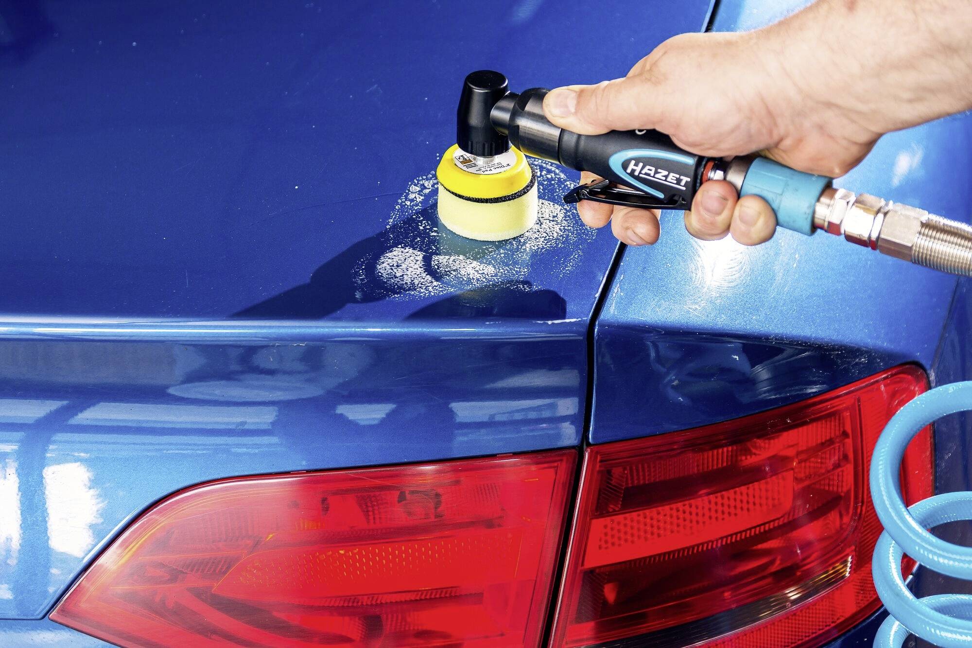 Rzemieślnik poleruje niebieski lakier samochodu elektrycznym urządzeniem polerskim. W tle czerwona lampa tylna.