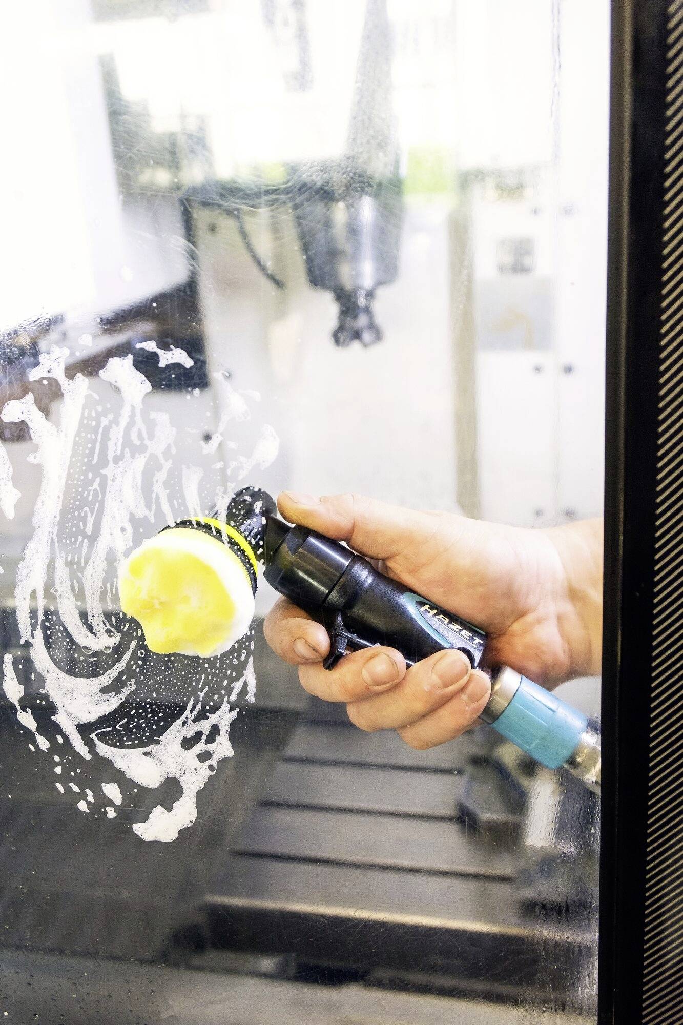 Dłoń czyści od wewnątrz szklane drzwi lub przezroczysty panel za pomocą środka czyszczącego i gąbki. W tle widać maszynę.