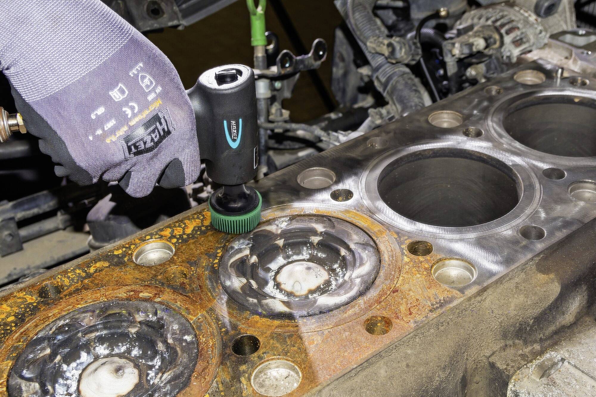 Dłoń w rękawicy trzyma narzędzie czyszczące nad zardzewiałą powierzchnią części samochodowych, usuwając plamy rdzy i brud.