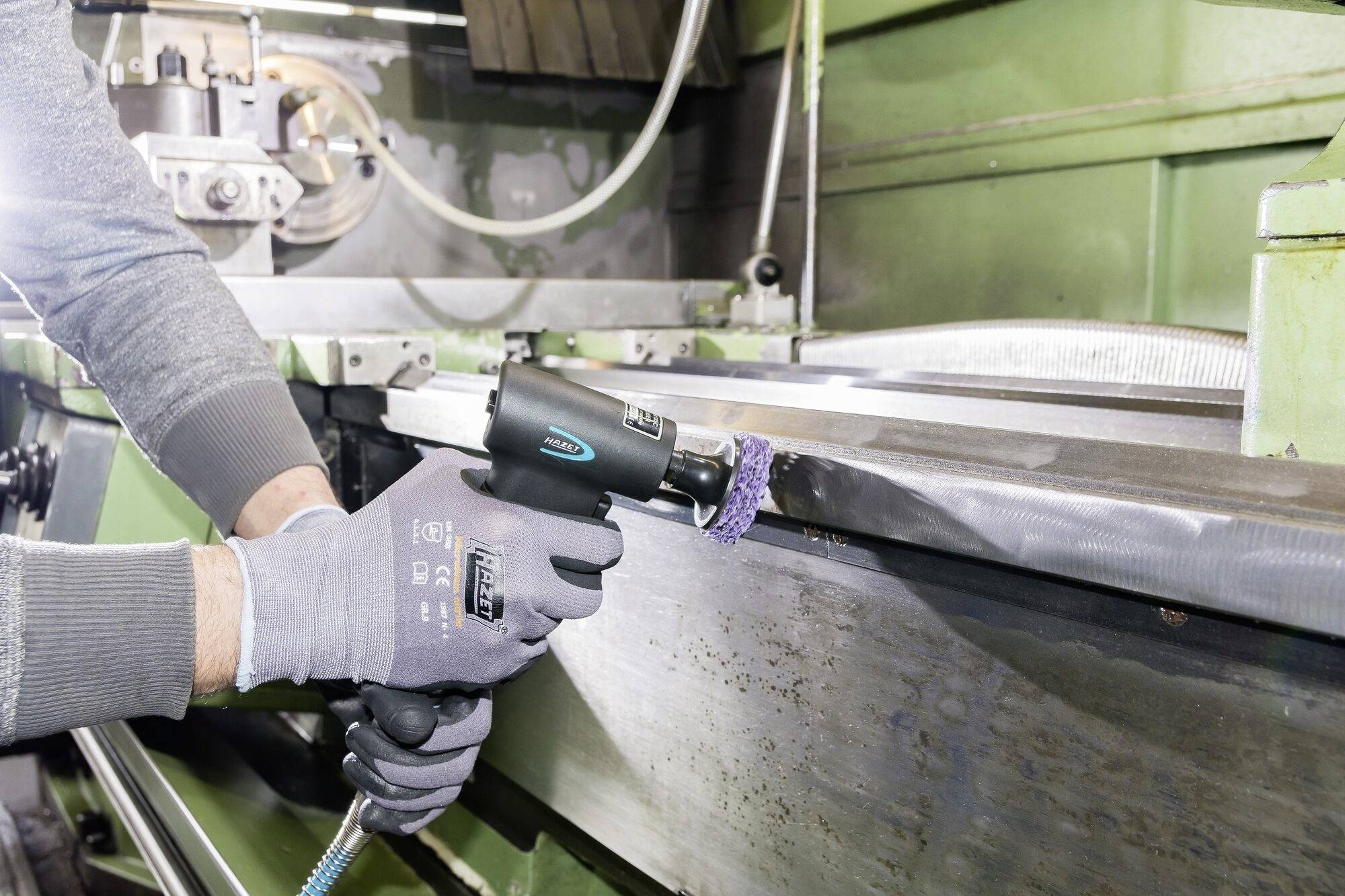 Osoba obrabia powierzchnię metalową ręcznym urządzeniem polerskim w warsztacie. Ma na sobie szare rękawice. Środowisko pracy.