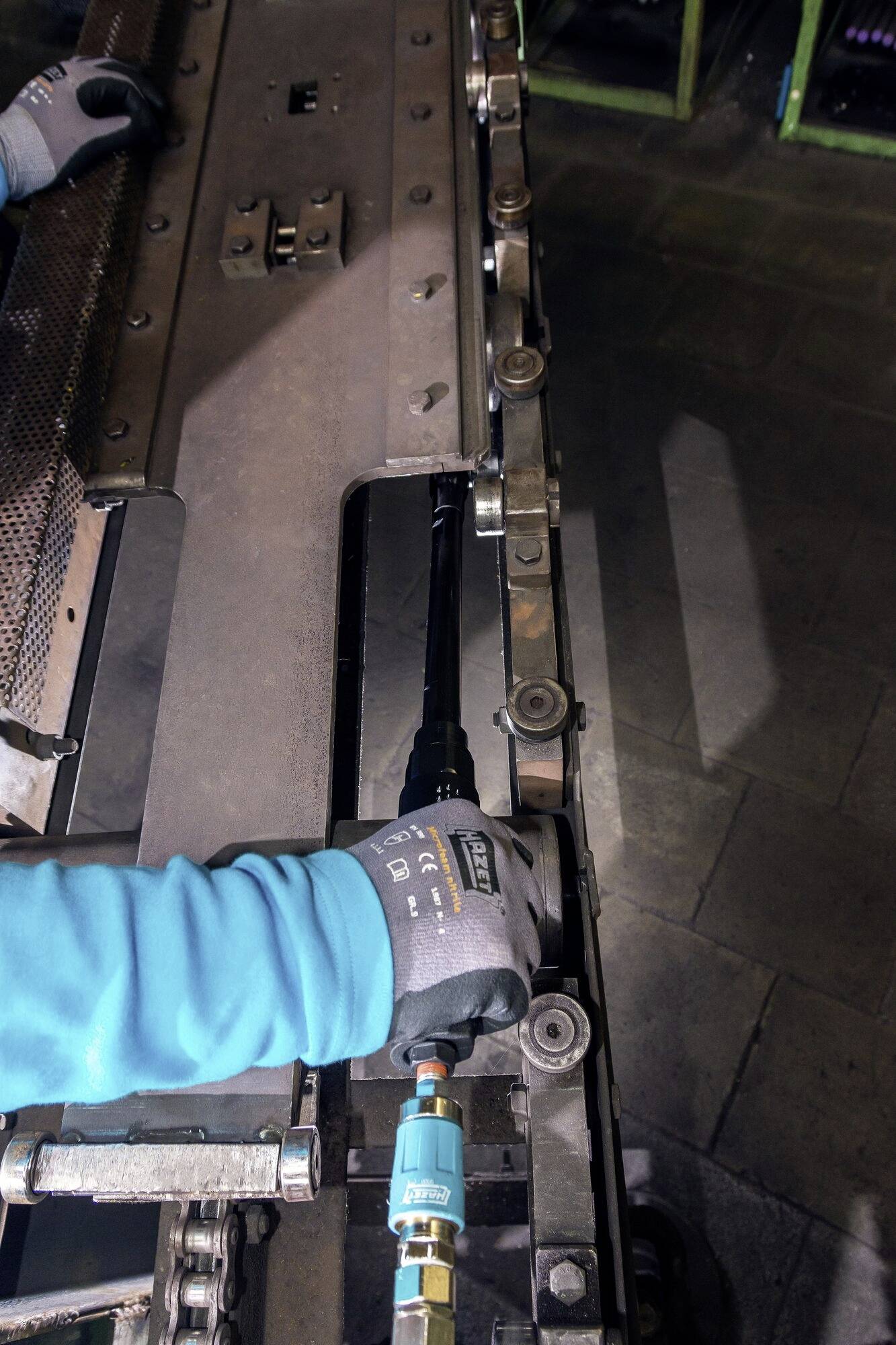 Pracownik w niebieskiej kurtce i szarych rękawiczkach naprawia dużą metalową maszynę, koncentrując się na mechanizmie.