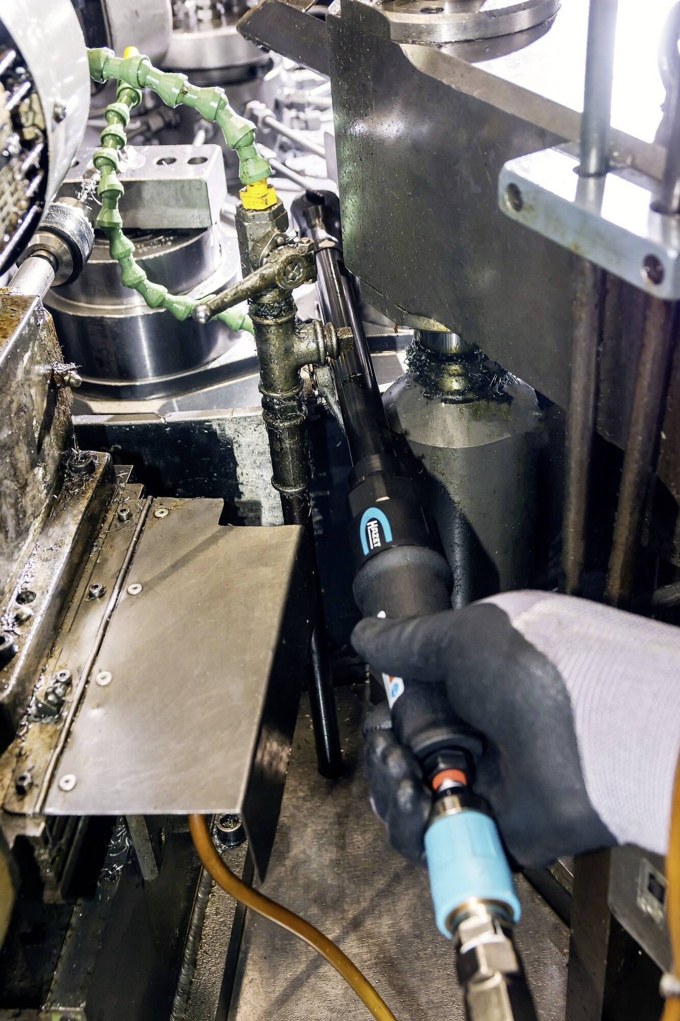 Osoba pracuje z narzędziem pneumatycznym pomiędzy metalowymi maszynami w środowisku warsztatowym. Podłączony jest przewód.