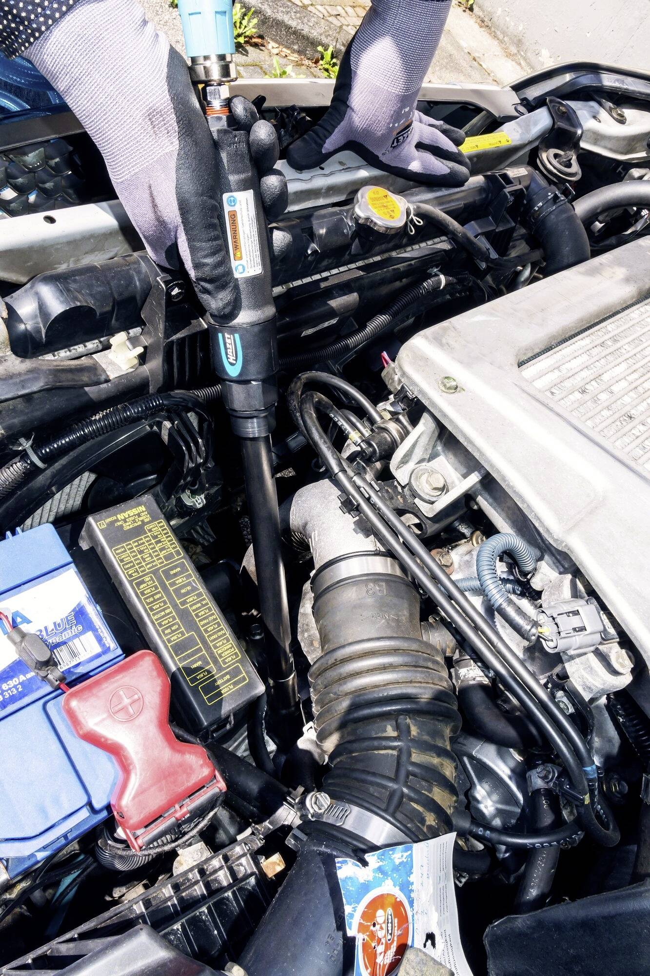 Osoba wymienia olej silnikowy w samochodzie. Pokrywa silnika jest otwarta, widoczne są części samochodowe i narzędzia.