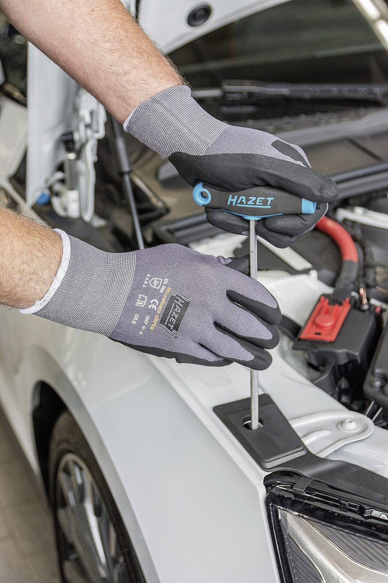 Osoba naprawia samochód śrubokrętem przed otwartą maską silnika. Ma na rękach szare rękawice.