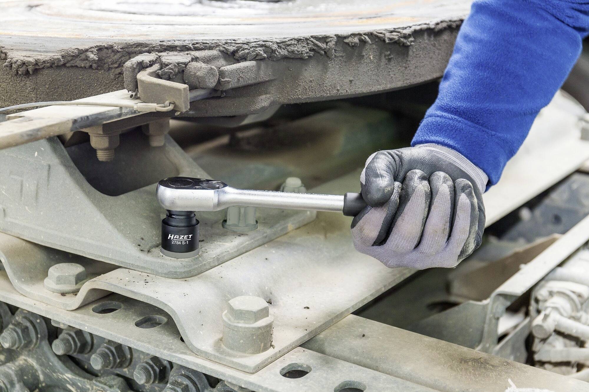 Osoba w niebieskich rękawach i czarnych rękawiczkach dokręca śrubę na dużej metalowej płycie za pomocą klucza zapadkowego.
