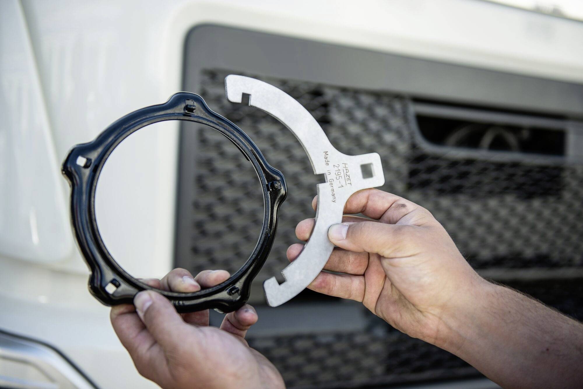 Dłonie trzymają części samochodowe przed kratką wlotu powietrza samochodu. Jedna z części jest czarna i okrągła, druga zaś metalowa.