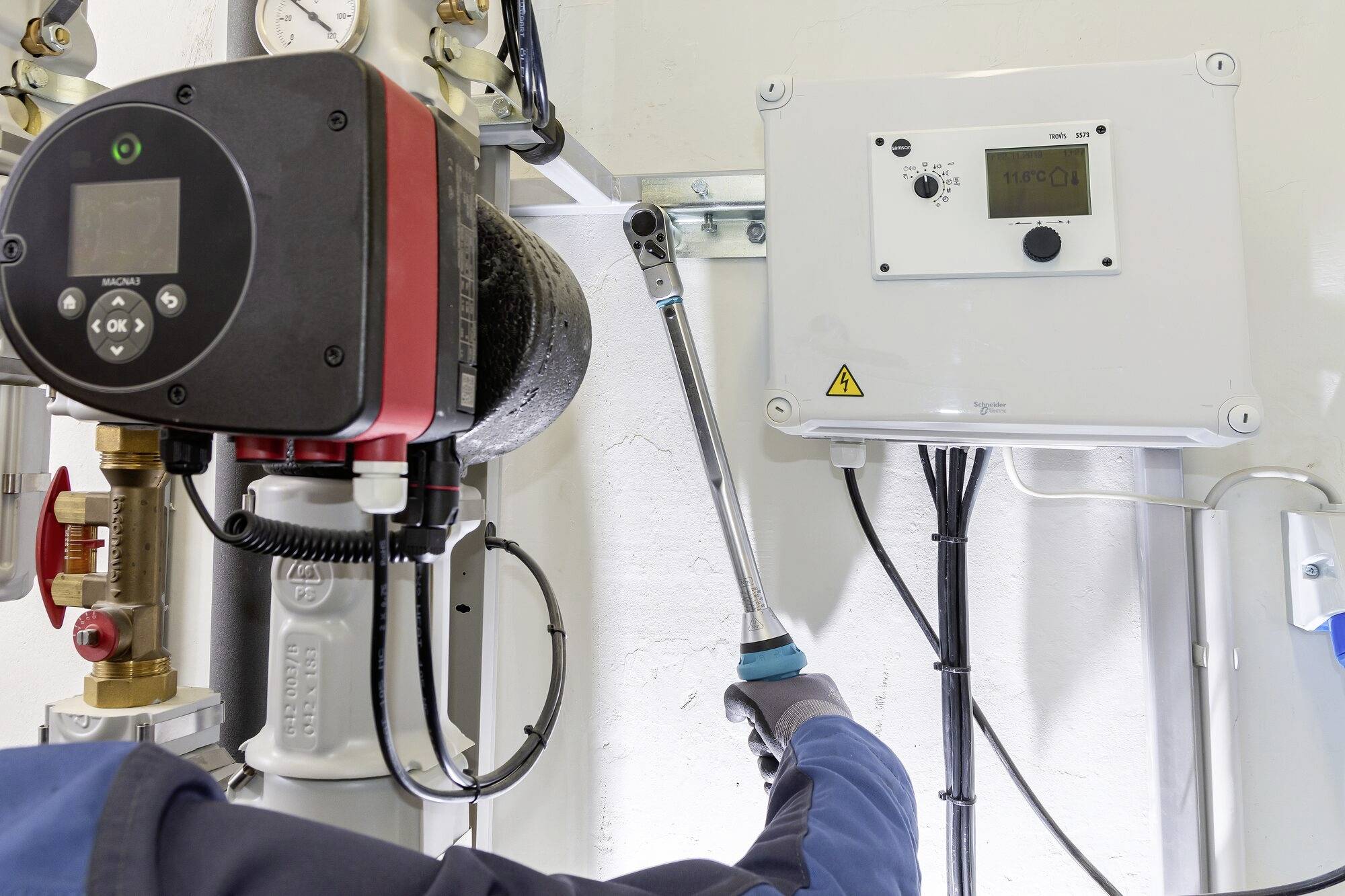 Osoba reguluje rurociąg w pomieszczeniu technicznym. Po lewej stronie duży czarny cylinder, po prawej biały panel sterowania.