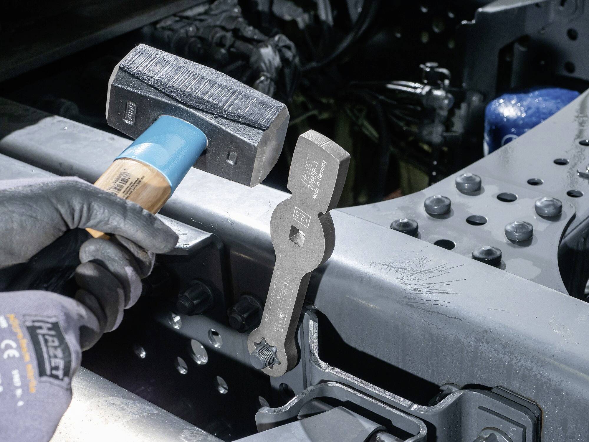 Mechanik używa specjalnego klucza i młotka do pracy przy ramie podwozia ciężarówki. Na pierwszym planie widoczne są części silnika.