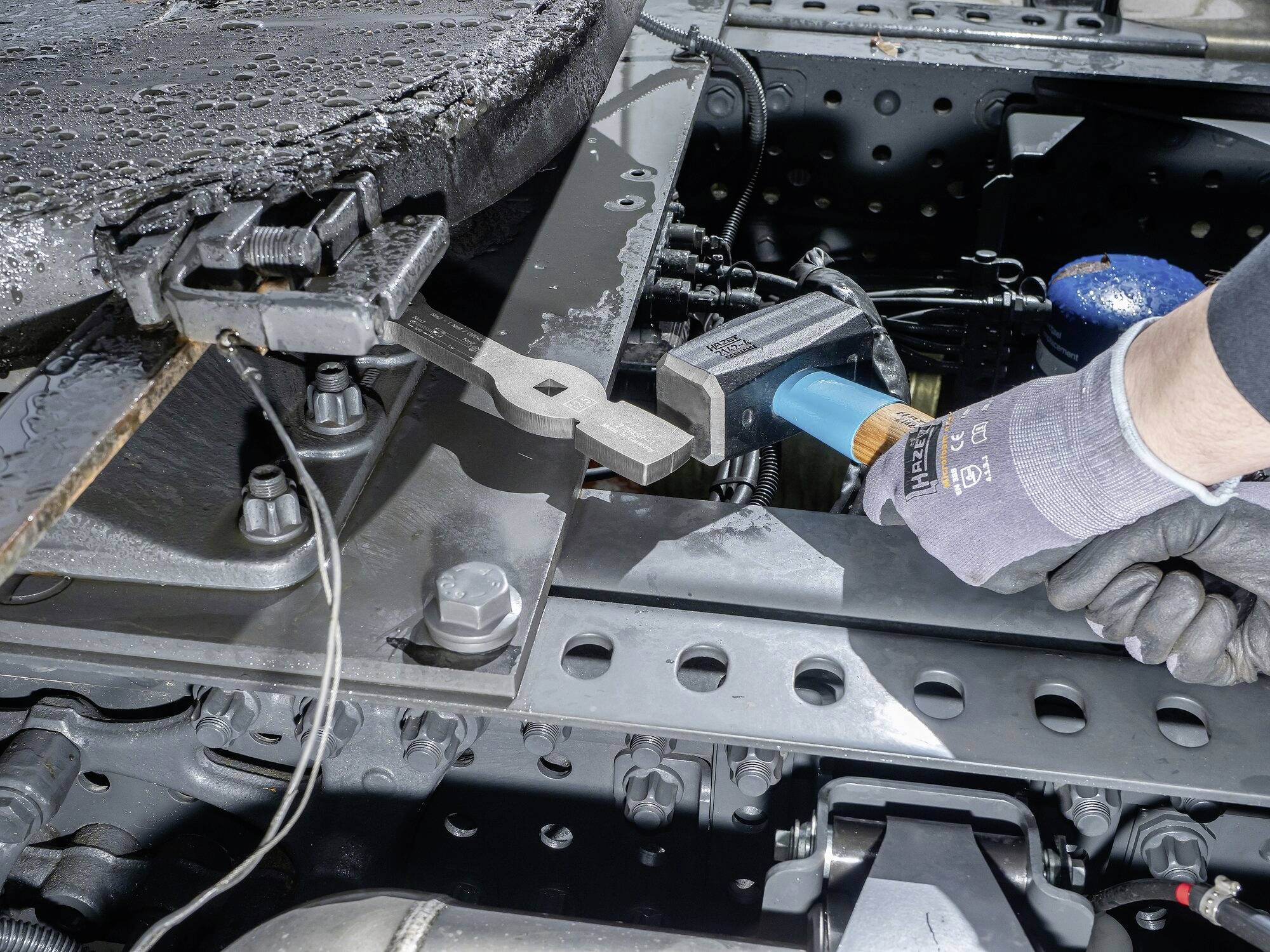 Osoba w rękawiczkach dokręca metalowy sworzeń na dużej ramie pojazdu. Wykonywane są prace konserwacyjne.