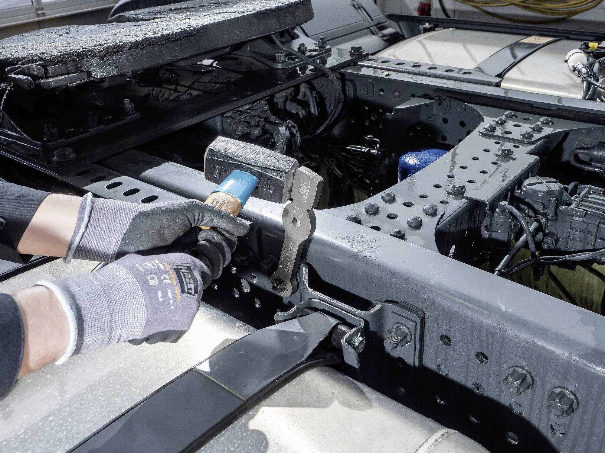 Osoba mocuje przyczepę do ramy samochodu ciężarowego za pomocą specjalnego narzędzia. Uwaga skupiona jest na elementach metalowych.