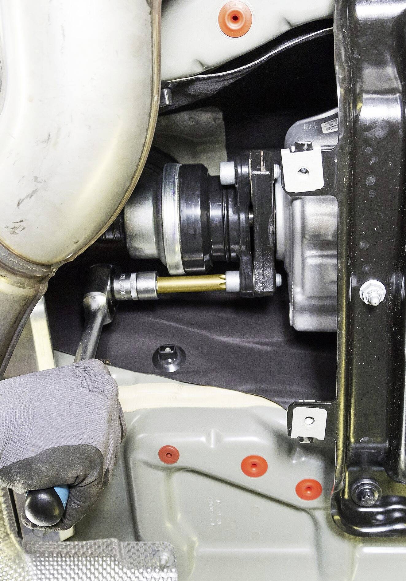 Osoba manipuluje kluczem płaskim przy pociąganiu dźwigni w warsztacie samochodowym, w pobliżu rury wydechowej oraz czarnego wspornika metalowego.