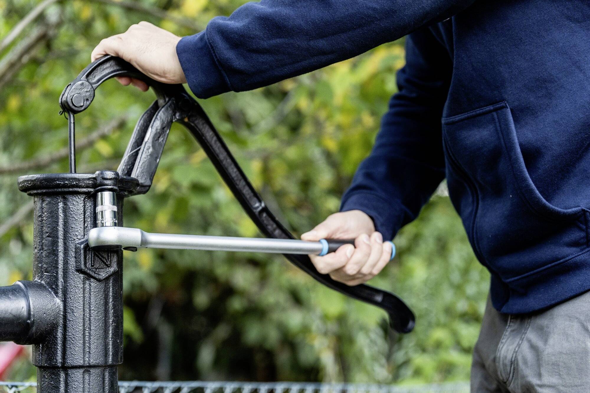 Osoba w niebieskim swetrze obsługuje ręczną pompę wodną na wolnym powietrzu.