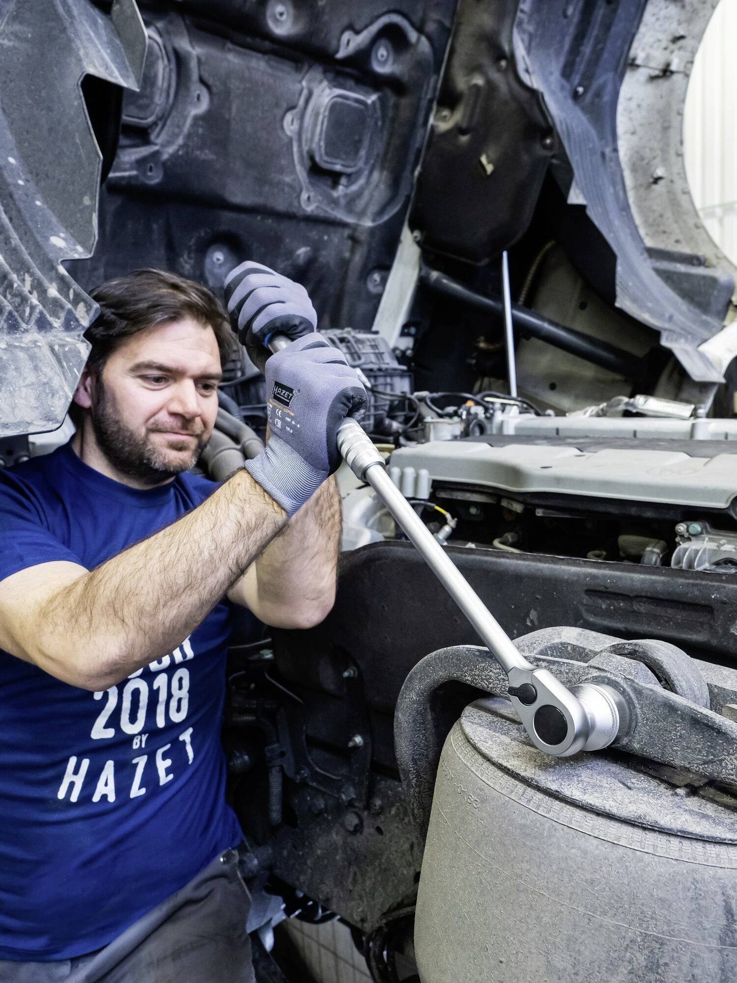 Mechanik pracujący przy pracy, dokręcający śrubę za pomocą grzechotki na silniku pojazdu. Ubrany w niebieskie ubranie robocze i rękawice.