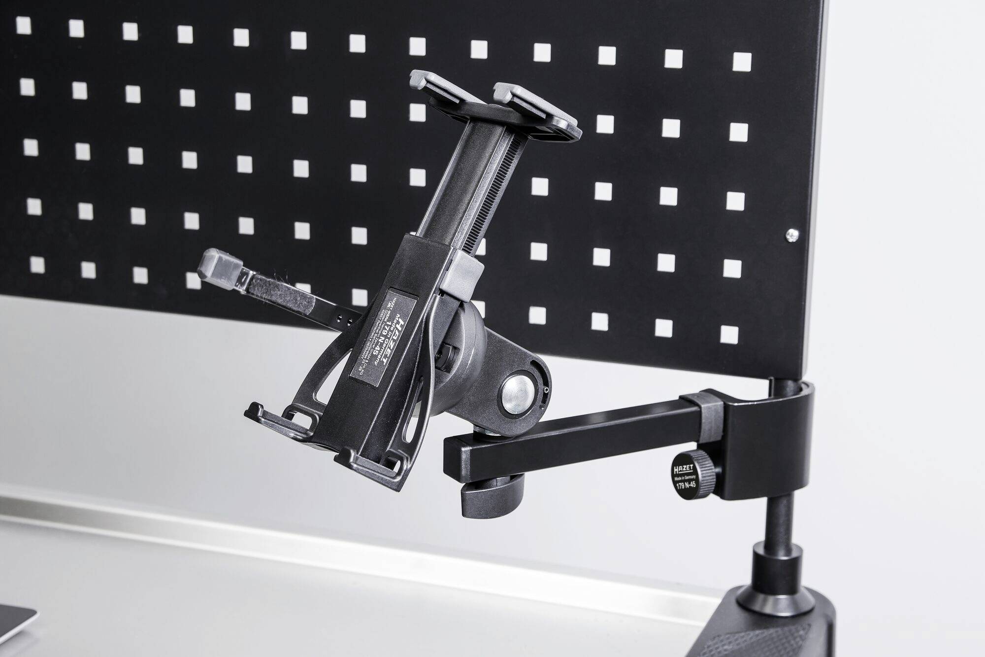 Czarny uchwyt na tablet z regulowanym ramieniem jest zamocowany na perforowanej ścianie i służy do elastycznego pozycjonowania urządzenia.