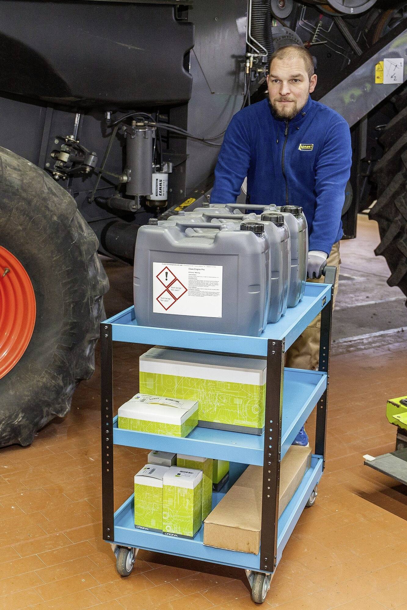 Mężczyzna przesuwa niebieski wózek z kanistrami paliwa obok dużych maszyn rolniczych.