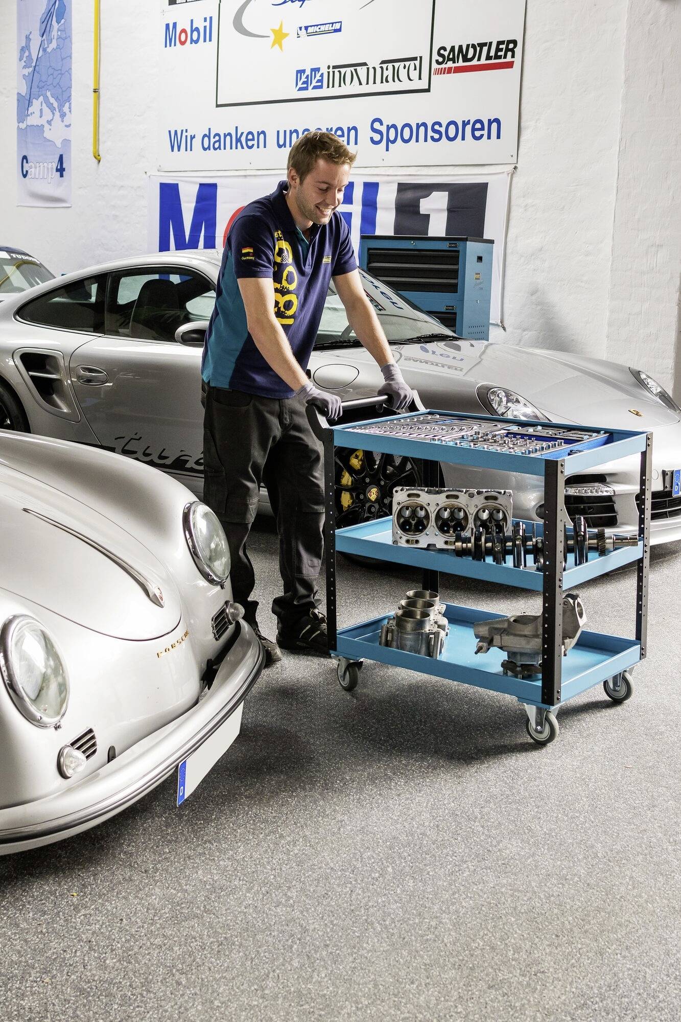 Mechanik stoi pomiędzy dwoma samochodami sportowymi w warsztacie; pracuje przy wózku narzędziowym pełnym części samochodowych.