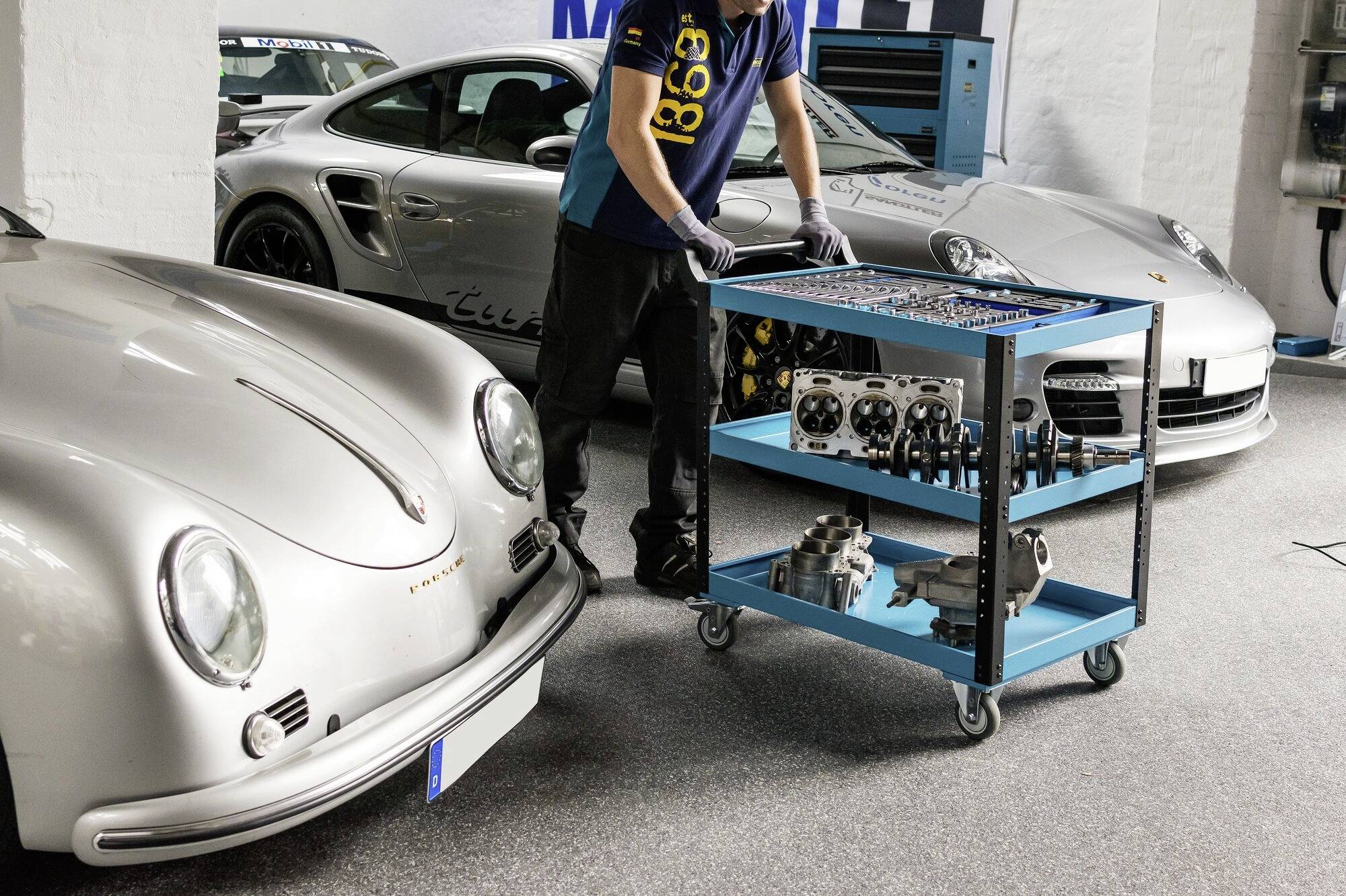 Pracownik warsztatu przesuwa wózek narzędziowy pomiędzy dwoma srebrnymi samochodami sportowymi. Na wózku znajdują się różne części samochodowe.