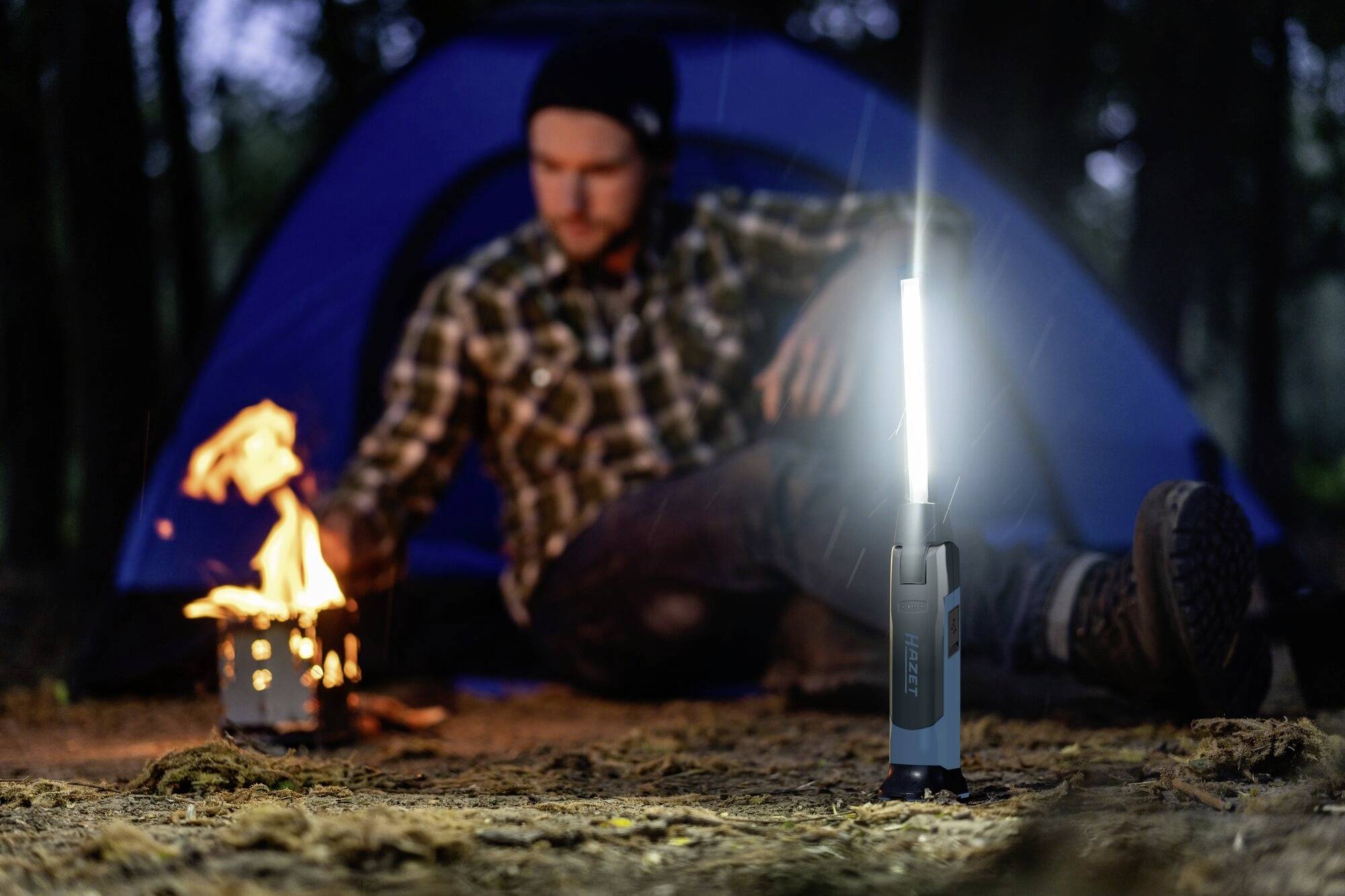 Mężczyzna siedzi w lesie przed namiotem i rozpala małe ognisko. Na pierwszym planie świeci lampa.