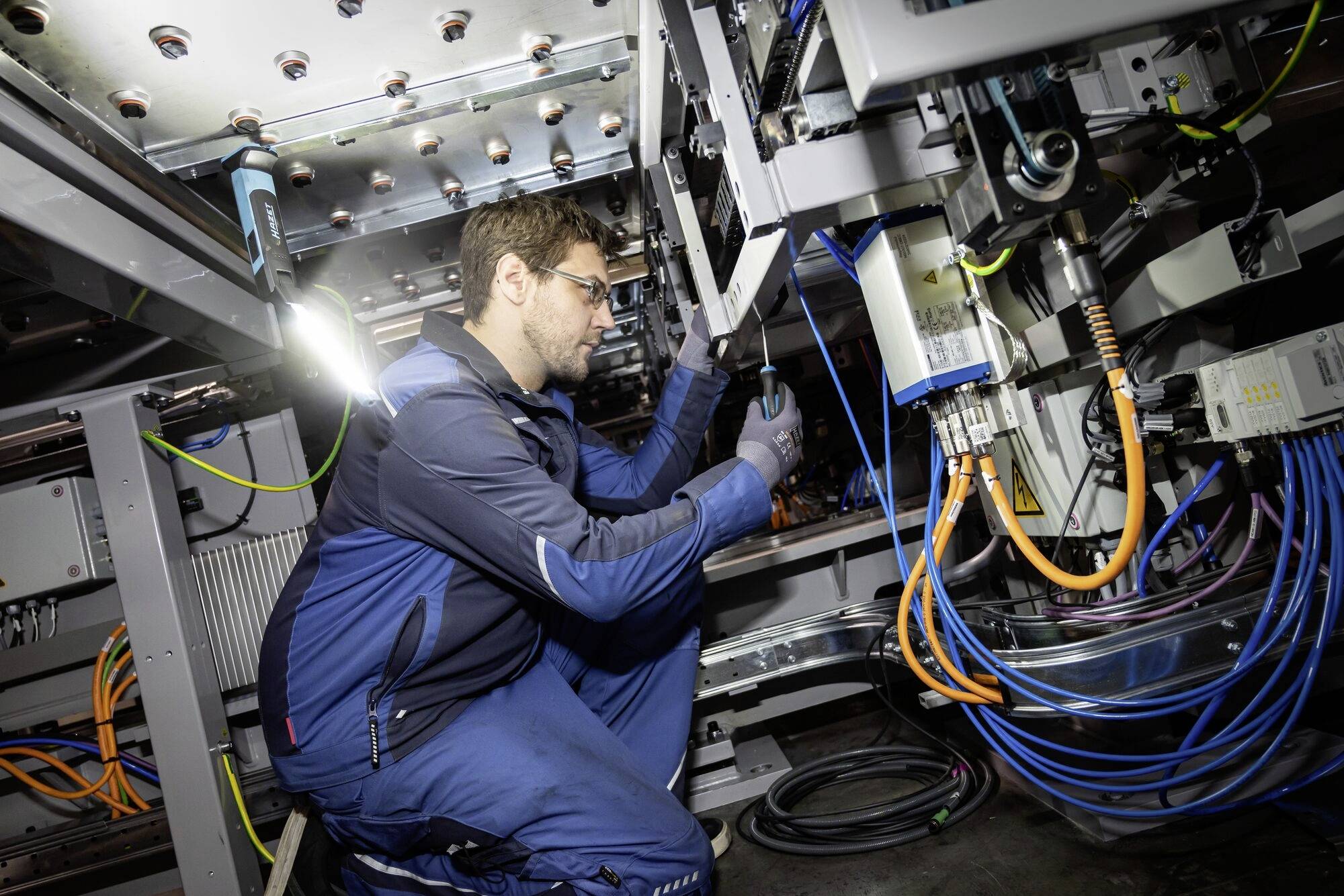 Technik w niebieskim ubraniu roboczym pracuje przy skomplikowanych maszynach z jaskrawo kolorowymi kablami w przemysłowym otoczeniu.