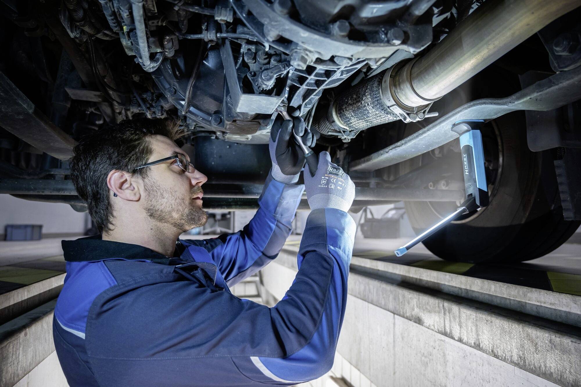 Mechanik w niebieskim kombinezonie roboczym pracuje pod pojazdem, sprawdza części za pomocą narzędzi i nosi rękawice ochronne.