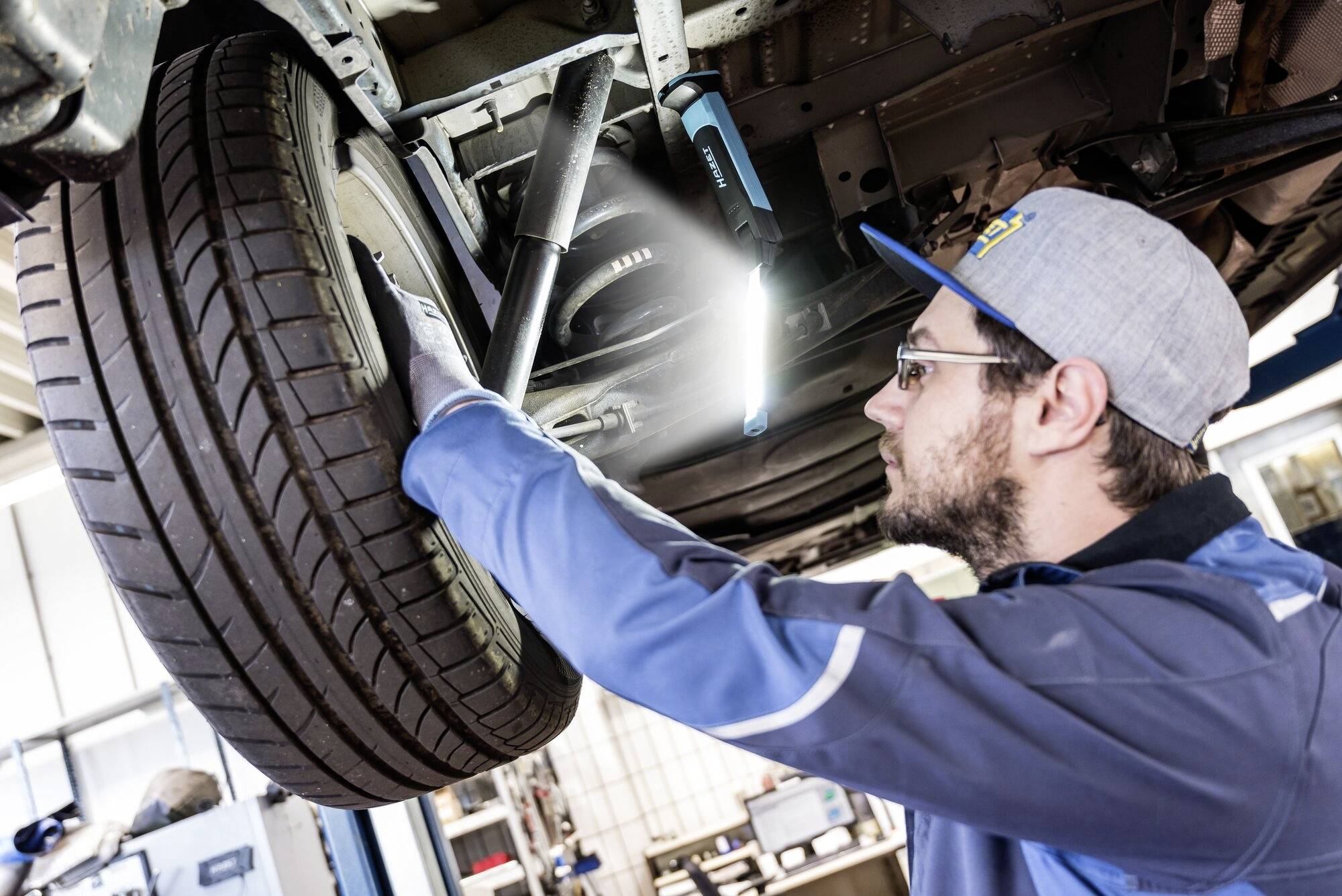 Mechanik naprawia samochód, który stoi na podnośniku. Sprawdza koło, używając źródła światła do dokładnej inspekcji.