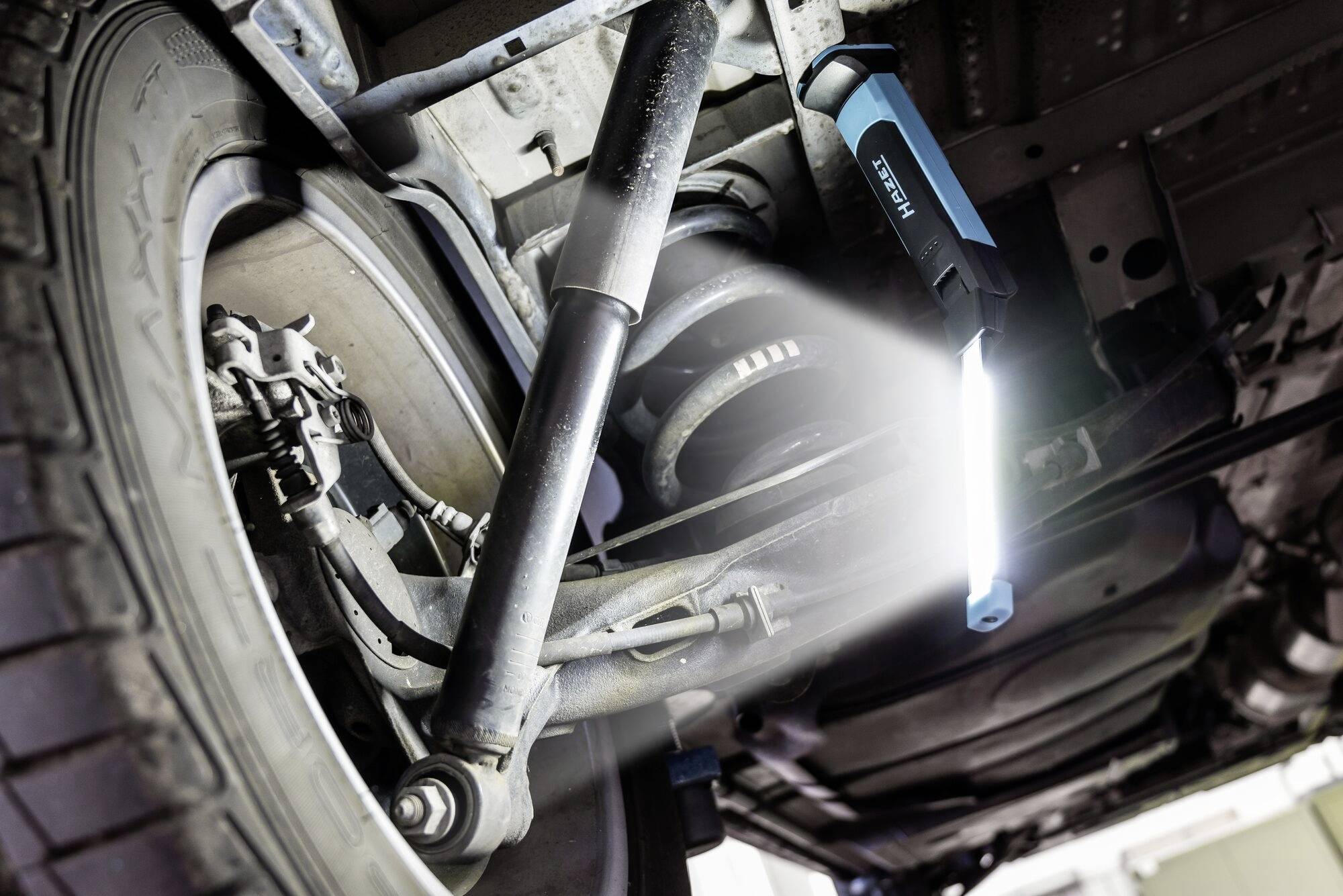 Widok spodu samochodu z oświetloną przestrzenią inspekcyjną, ze szczególnym naciskiem na zawieszenie i amortyzatory. Widoczne szczegóły mechaniczne.