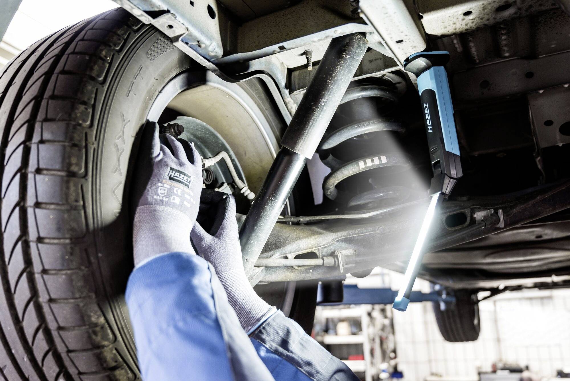 Osoba naprawia samochód, używając specjalistycznych narzędzi. Fokus jest skierowany na oponie i zawieszeniu pojazdu.