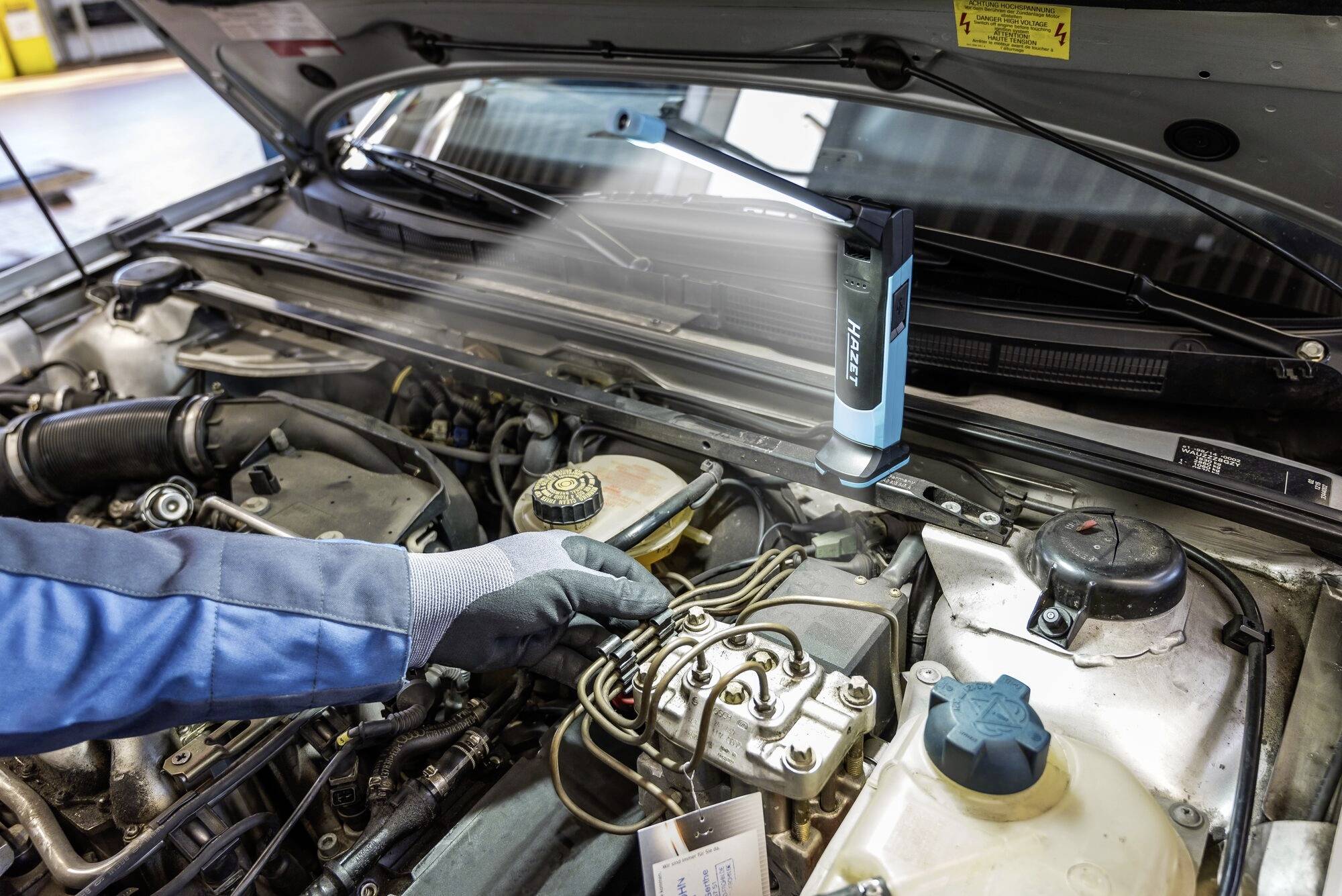 Osoba ubrana w niebieską kurtkę i w rękawiczkach kontroluje komorę silnika samochodu. Latarka oświetla badany obszar.