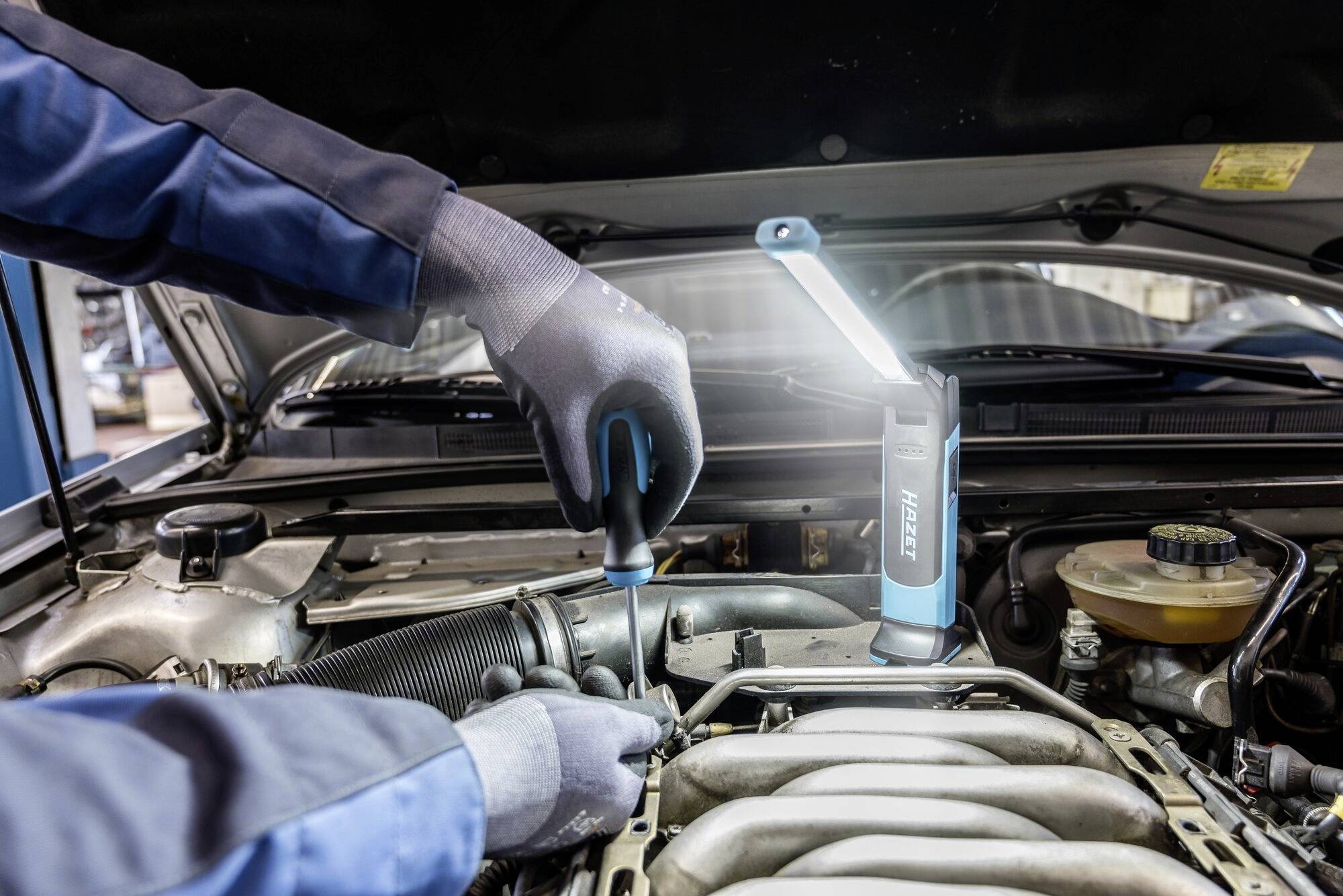 Mechanik naprawia silnik narzędziem, oświetlony przenośną lampą. Fokus na rzemieślniczą pracę w komorze silnika samochodu.