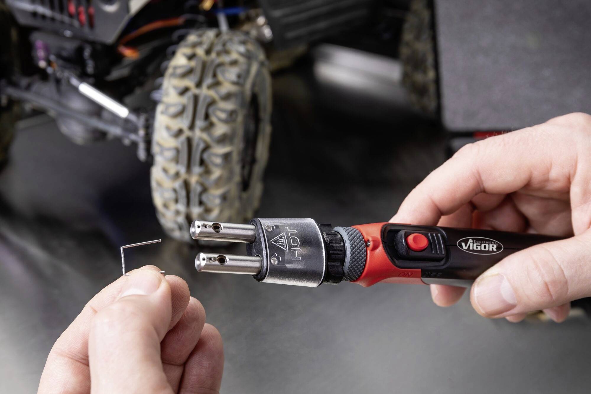 Osoba trzyma czerwony lutownik i małą wygiętą drucianą część. W tle widoczne są duże opony pojazdu.