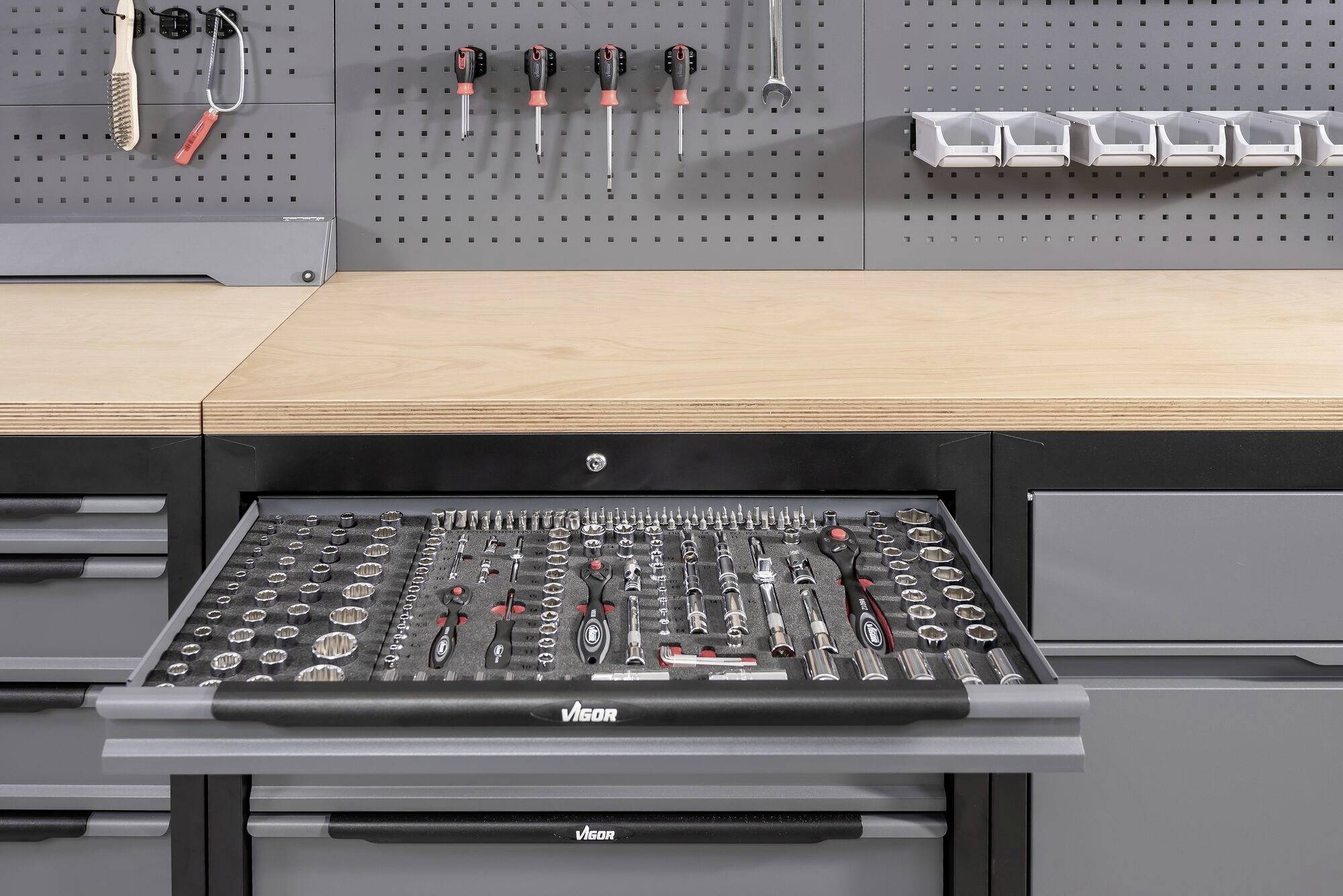 Otwarta szuflada warsztatowa prezentuje różne narzędzia, w tym śrubokręty i klucze nasadowe. W tle: szara ściana z hakami.