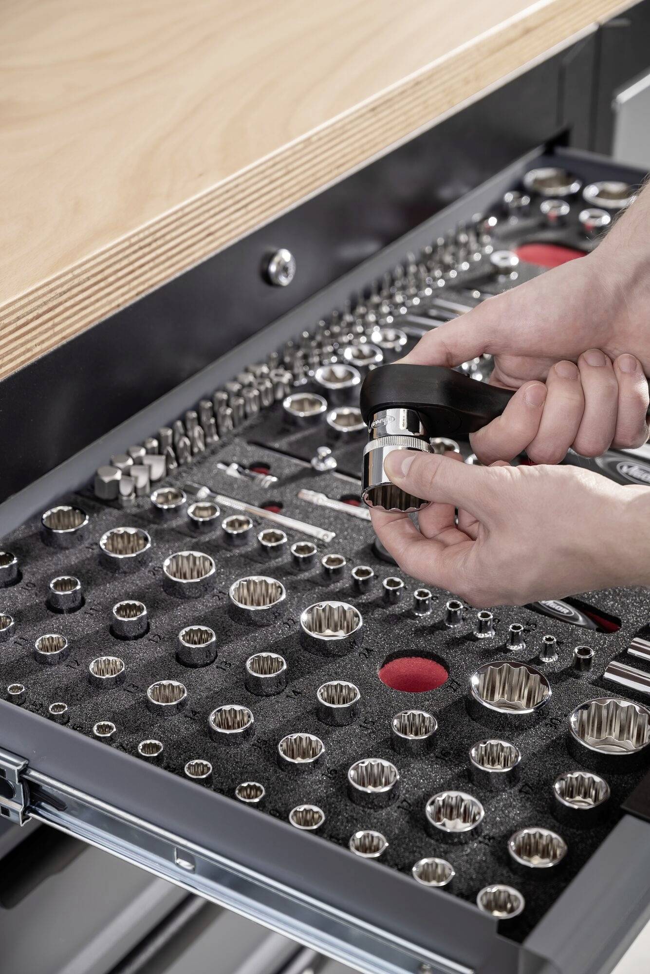 Otwarta szuflada z narzędziami z różnymi kluczami nasadowymi i parą rąk chwytających klucz zapadkowy.