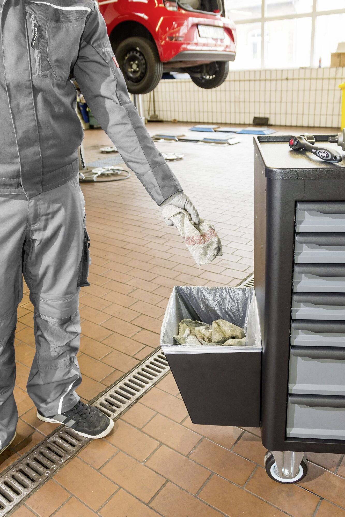 Osoba wrzuca szmatkę do kosza na śmieci obok wózka narzędziowego w warsztacie samochodowym. W tle widać samochód uniesiony na podnośniku.