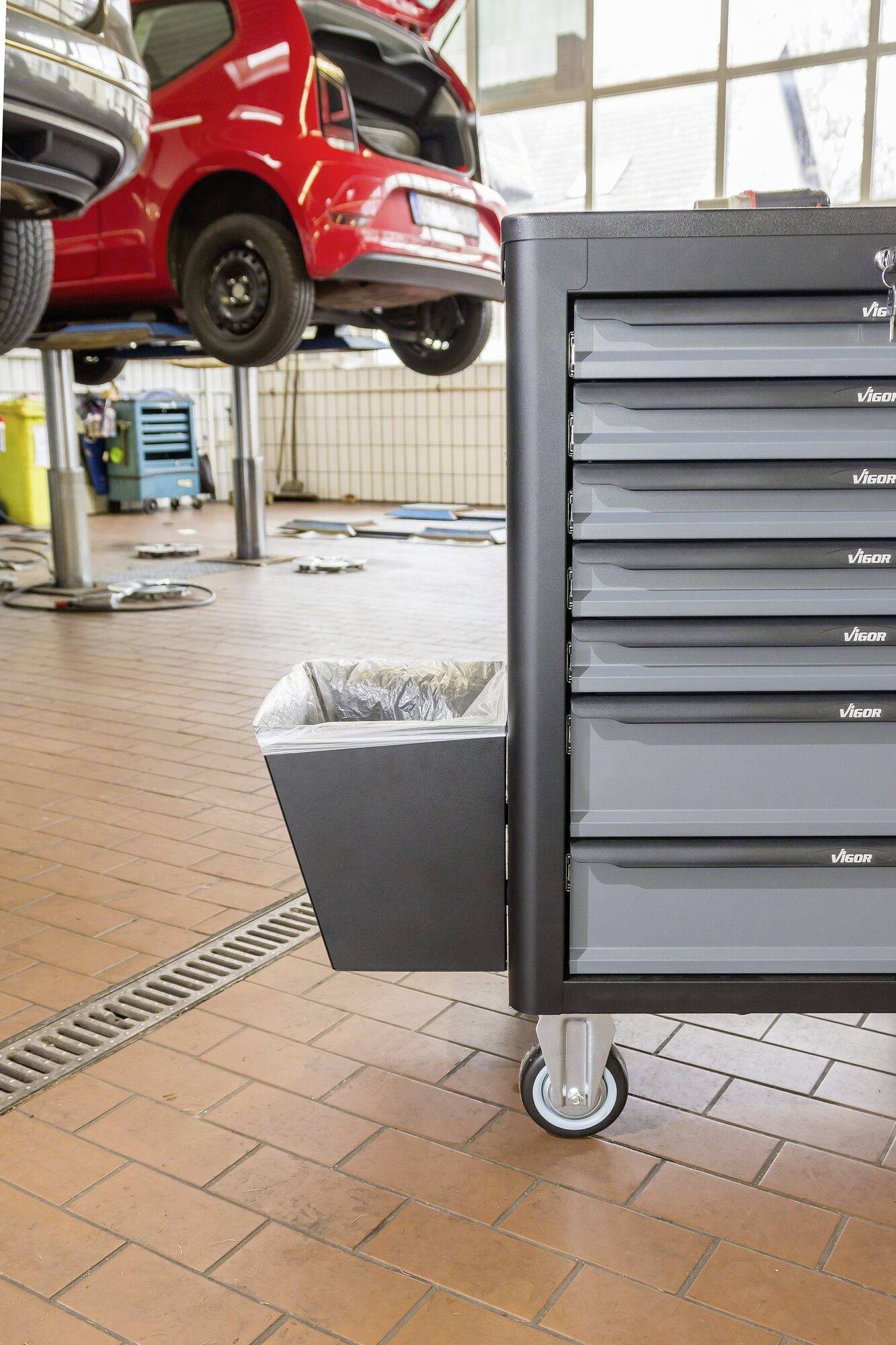 Warsztat z czerwonymi samochodami na podnośnikach, duży wózek warsztatowy z szufladami i kosz na odpady w pierwszym planie.