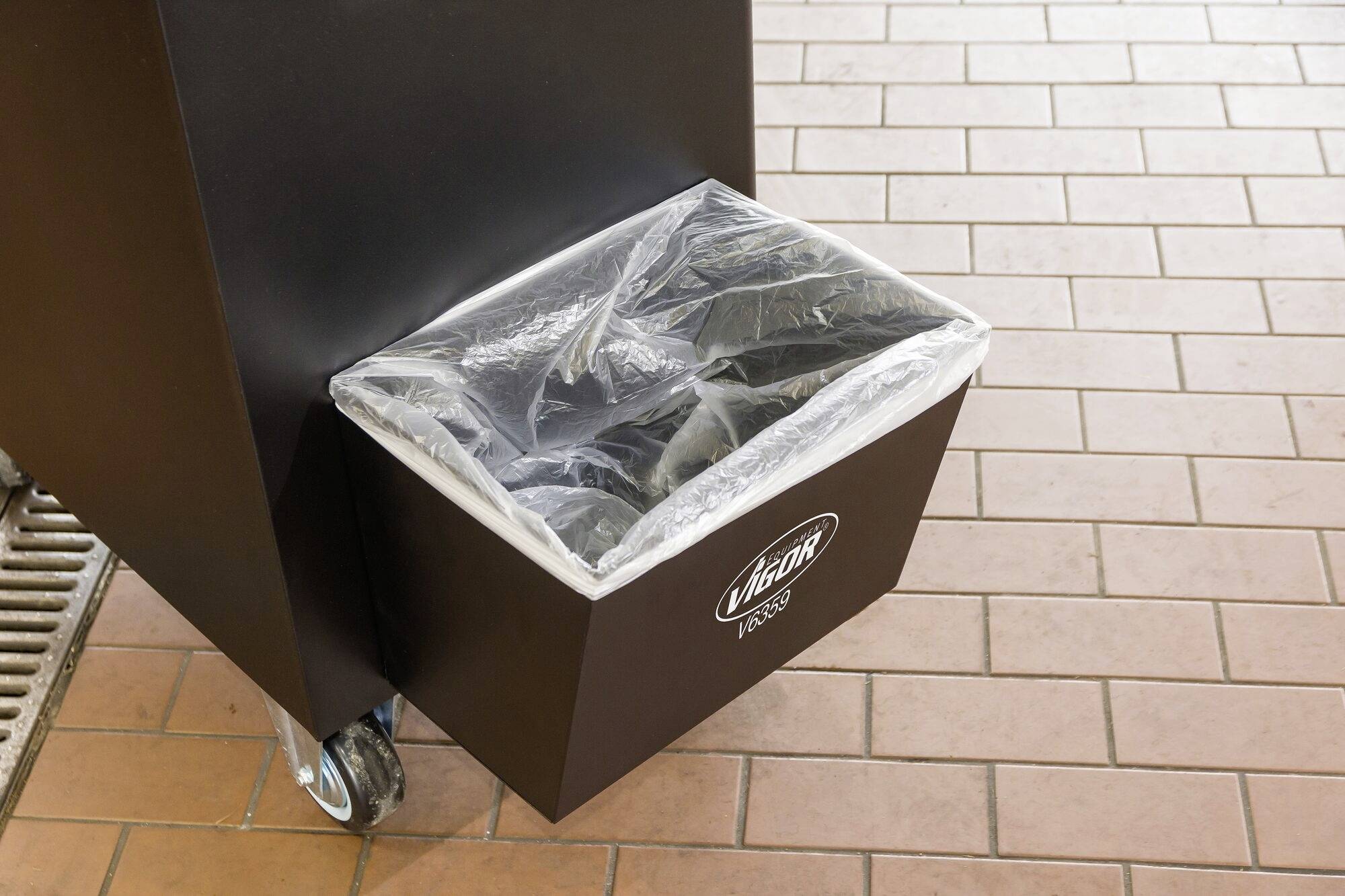 Czarny kosz na śmieci z przezroczystą plastikową torbą, stojący na podłodze wyłożonej płytkami.